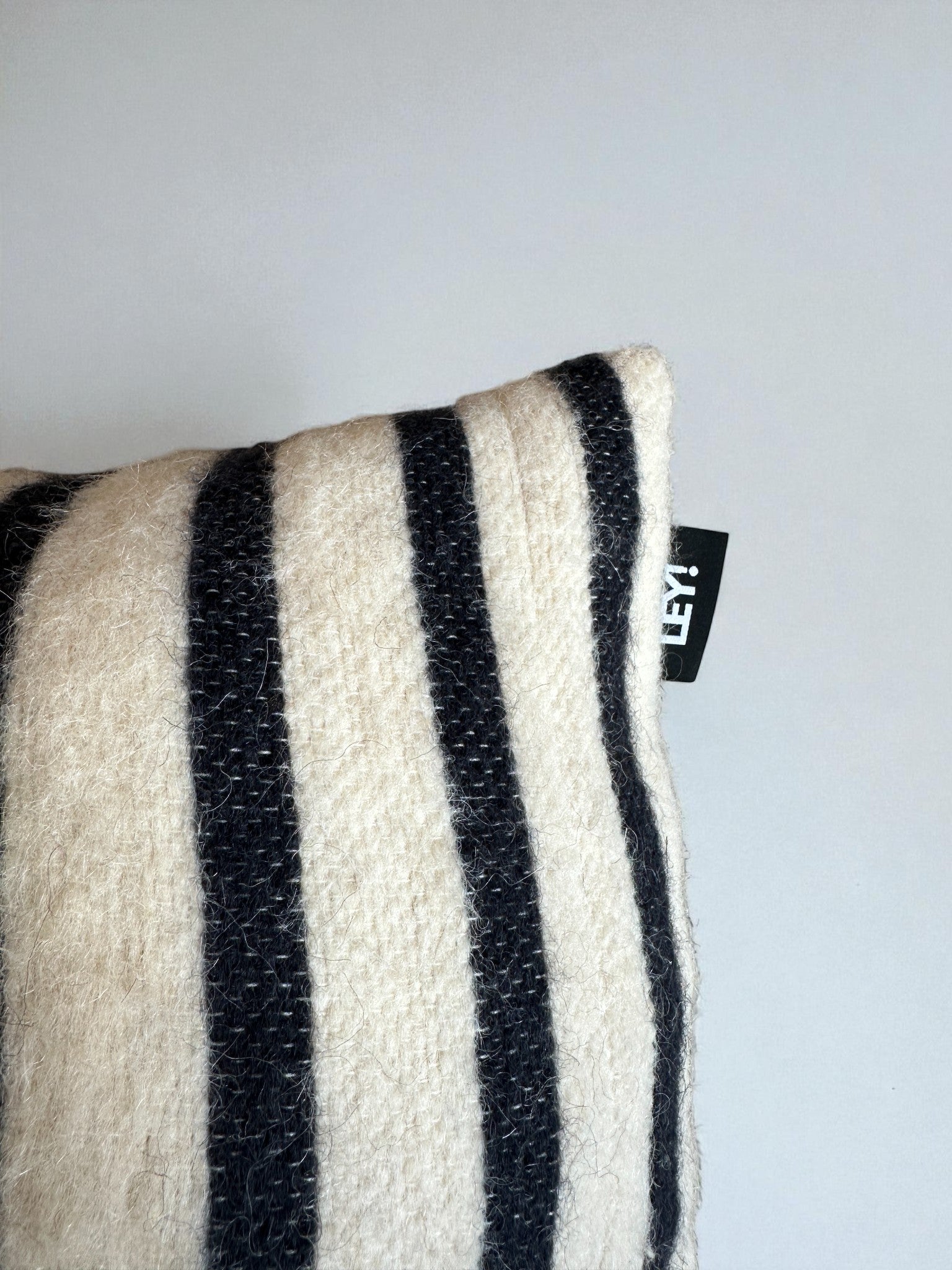 Eine Nahaufnahme des weichen, rechteckigen LEY! Kissen - WINTER EDITION - TAU - 30 x 50, mit schwarzen und cremefarbenen vertikalen Streifen und einem kleinen schwarzen Etikett mit weißem Text an der Seite, vor einem schlichten hellgrauen Hintergrund.