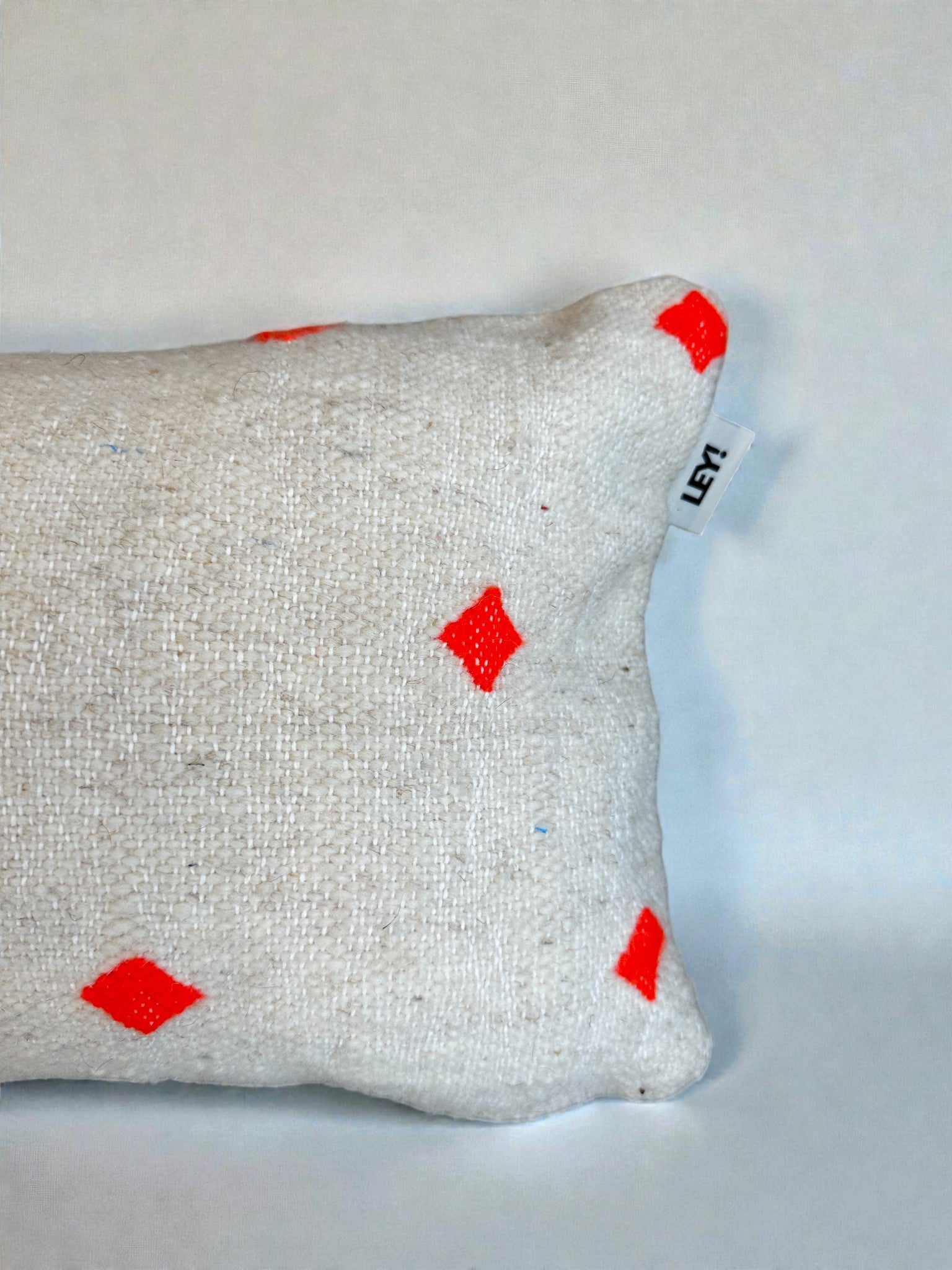 Das LEY! Kissen - Mona Orange (30x50cm) ist ein beigefarbener, handgefertigter Schurwoll-Kissenbezug mit strukturiertem Stoff und roten Rauten, der ein kleines weißes Schildchen mit schwarzem Text am Rand auf weißem Hintergrund aufweist.