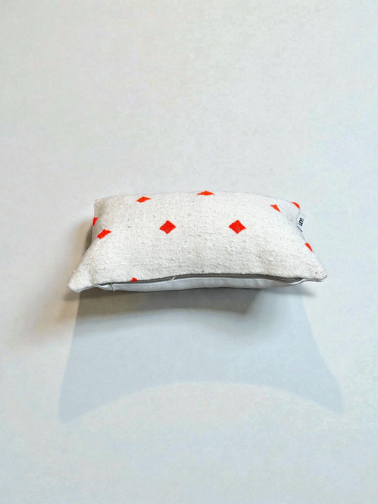Ein kleines rechteckiges LEY! Kissen - Mona Orange - 30 x 50 mit einem hellgrauen Bezug, auf dem rote Rauten verstreut sind, ruht auf einer schlichten weißen Fläche und wirft einen weichen Schatten.
