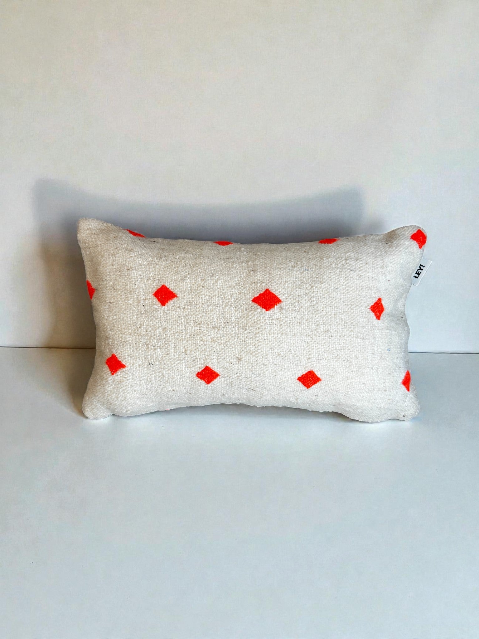 Ein rechteckiges weißes Kissen - Mona Orange - 30x50cm von LEY! mit kleinen roten Rauten, die aufrecht auf einem schlichten weißen Hintergrund stehen. Mit Schurwolle-Kissenbezug in Handarbeit im Marrakesch-Stil für eine handwerkliche Note.