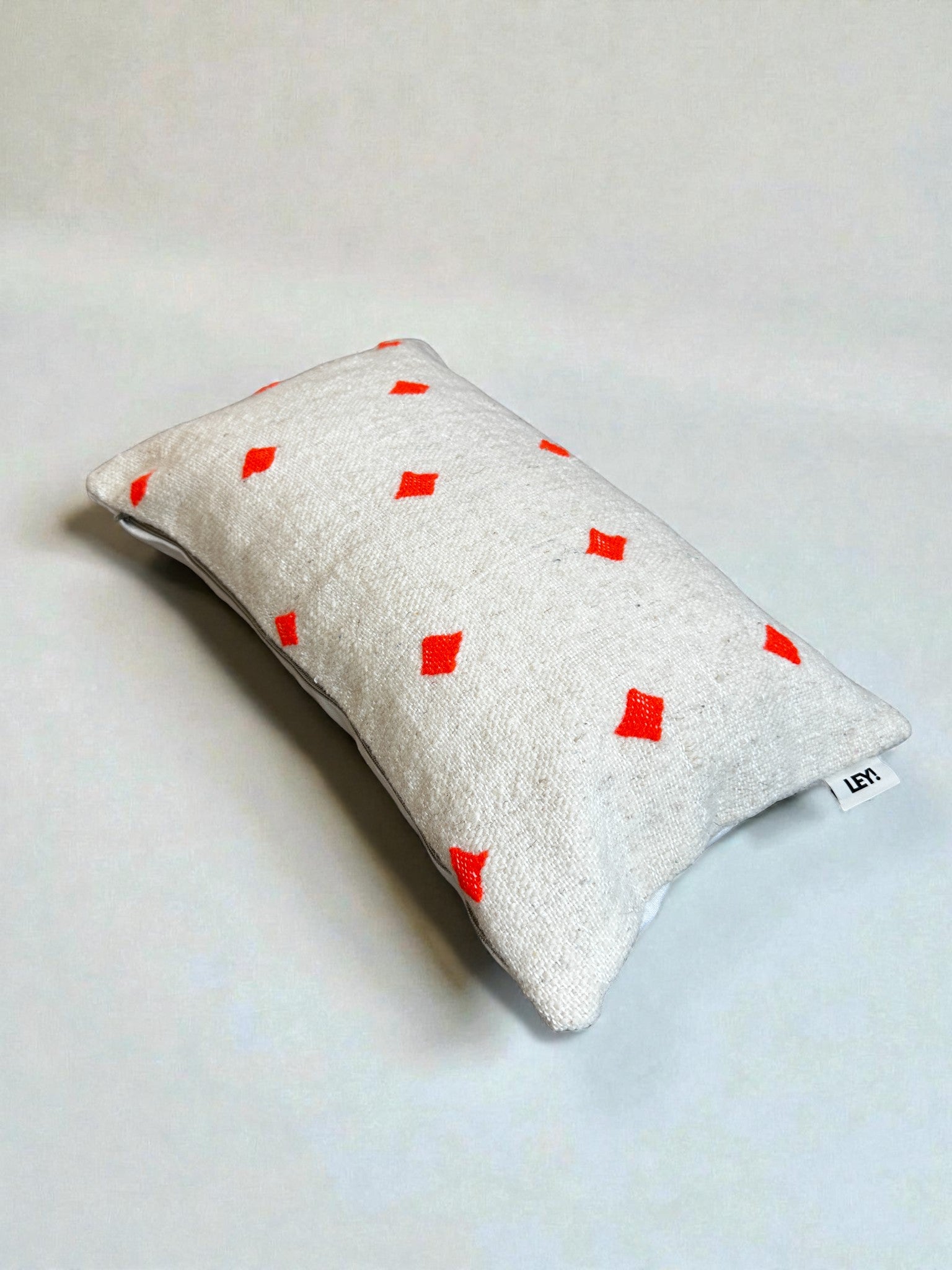 Das Kissen - Mona Orange - 40 x 60 von LEY! hat einen rechteckigen weißen Bezug mit roten Rautenmotiven, Handarbeit im Marrakesch-Stil in Schurwolle und einem kleinen Stoffanhänger, der auf einem schlichten, hellen Hintergrund angebracht ist.