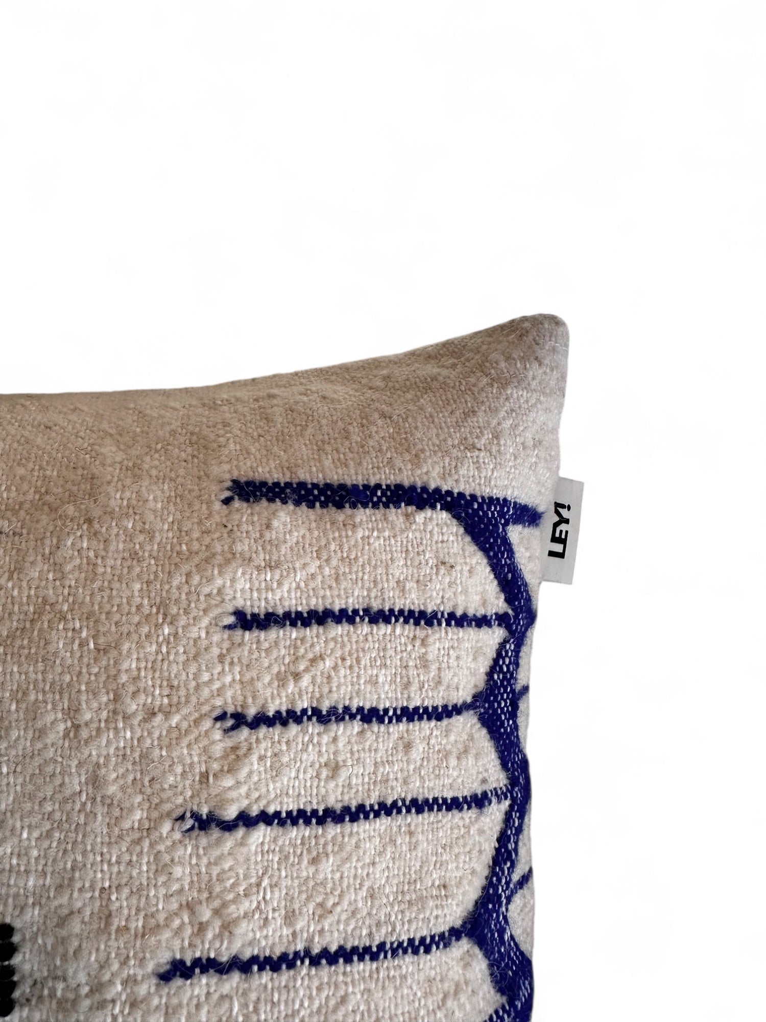 Eine Nahaufnahme des LEY! Kissen - Colorful Line - 40 x 60 zeigt cremefarbenen Stoff mit blauer geometrischer Stickerei und ein kleines weißes LEY!-Tag auf der oberen rechten Naht vor einem schlichten weißen Hintergrund.