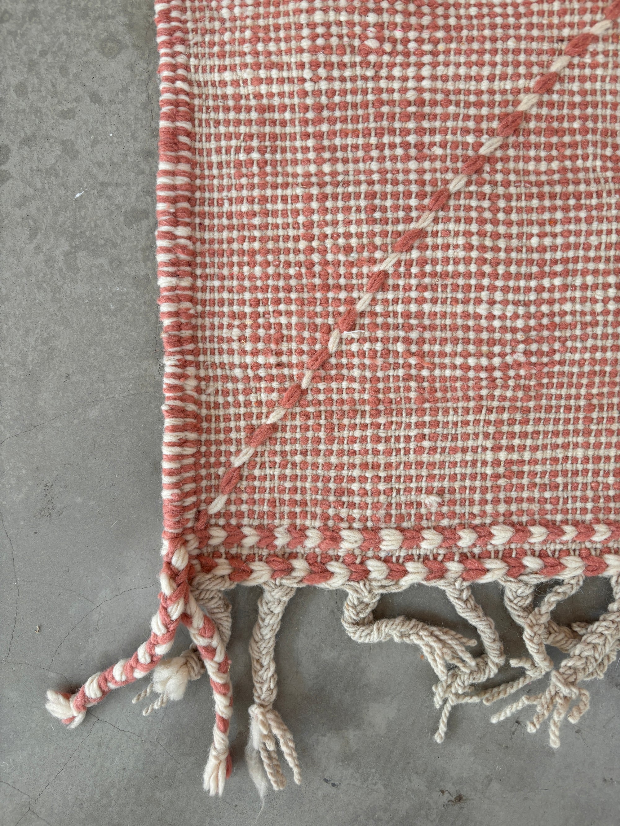 Eine Nahaufnahme des LEY Teppich - Ava (0,85 x 3,00) von LEY! mit einem rot-weiß karierten Flachgewebe-Muster mit cremefarbenen Quasten und einer gedrehten Kordel, ausgestellt auf einem grauen Betonboden.