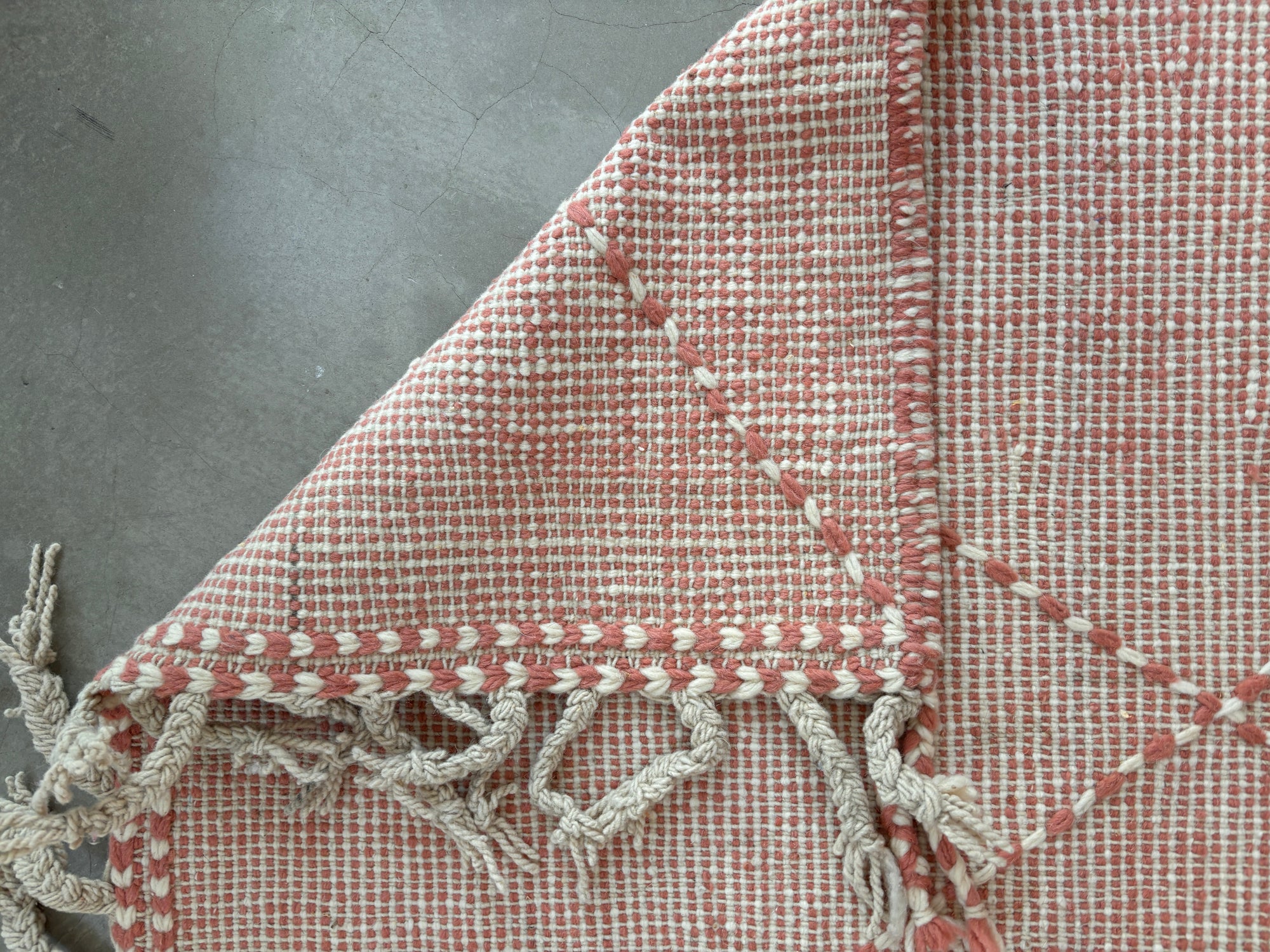 Eine Nahaufnahme des LEY Teppichs - Ava (0,85 x 3,00) in rosa und weiß von LEY! auf einem grauen Boden mit einer gefalteten Ecke, die beide Seiten zeigt, und mit Fransen, die den einzigartigen marokkanisch inspirierten Charme hervorheben.
