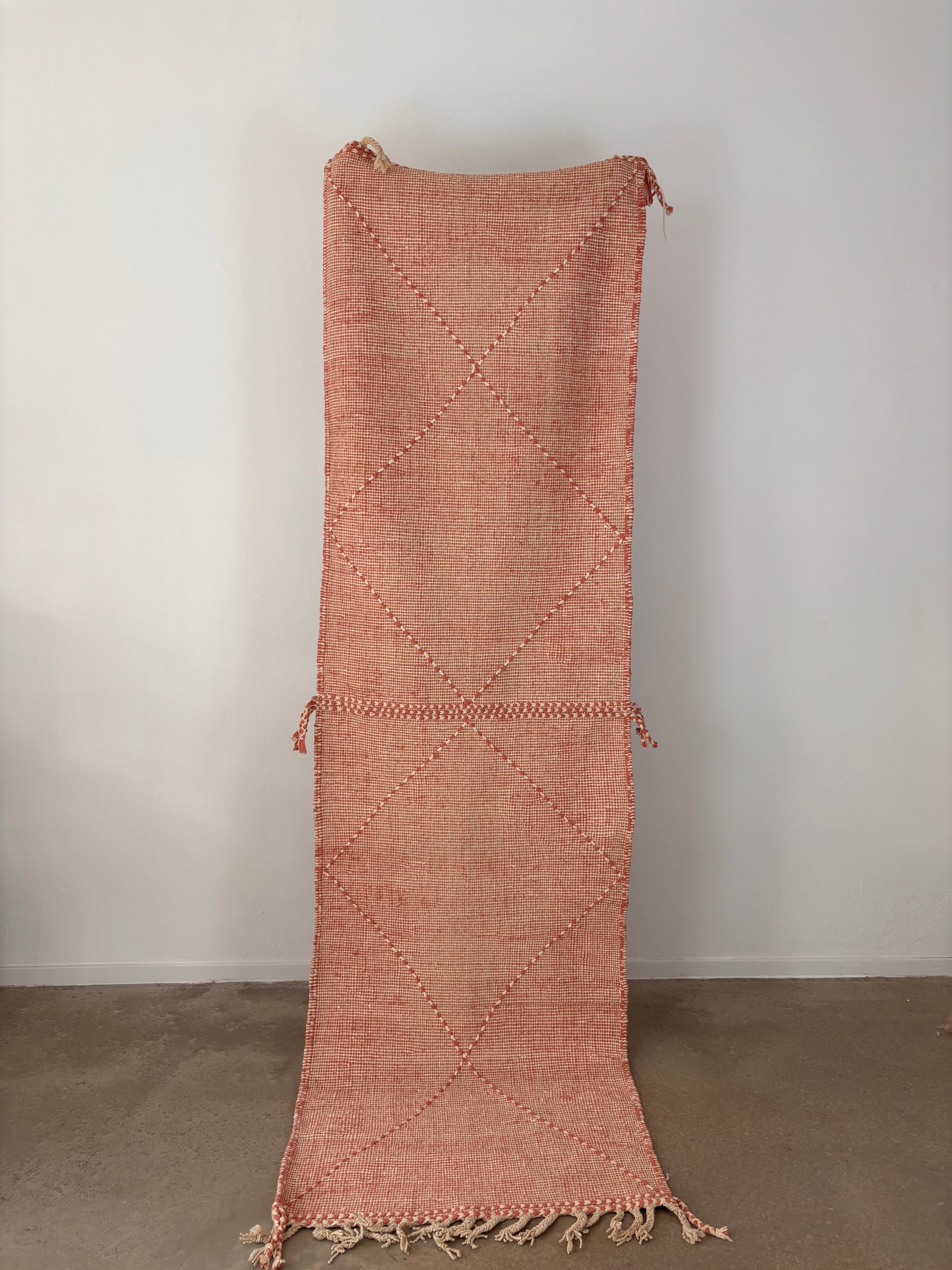 Der LEY! Teppich - Ava - 0,85 x 3,00 ist ein handgeknüpfter Kelim Läufer in Rot und Beige mit einem Rautenmuster, Quasten an einem Ende und ausgefransten Kanten, der vertikal vor einer weißen Wand und einem Betonboden gezeigt wird.