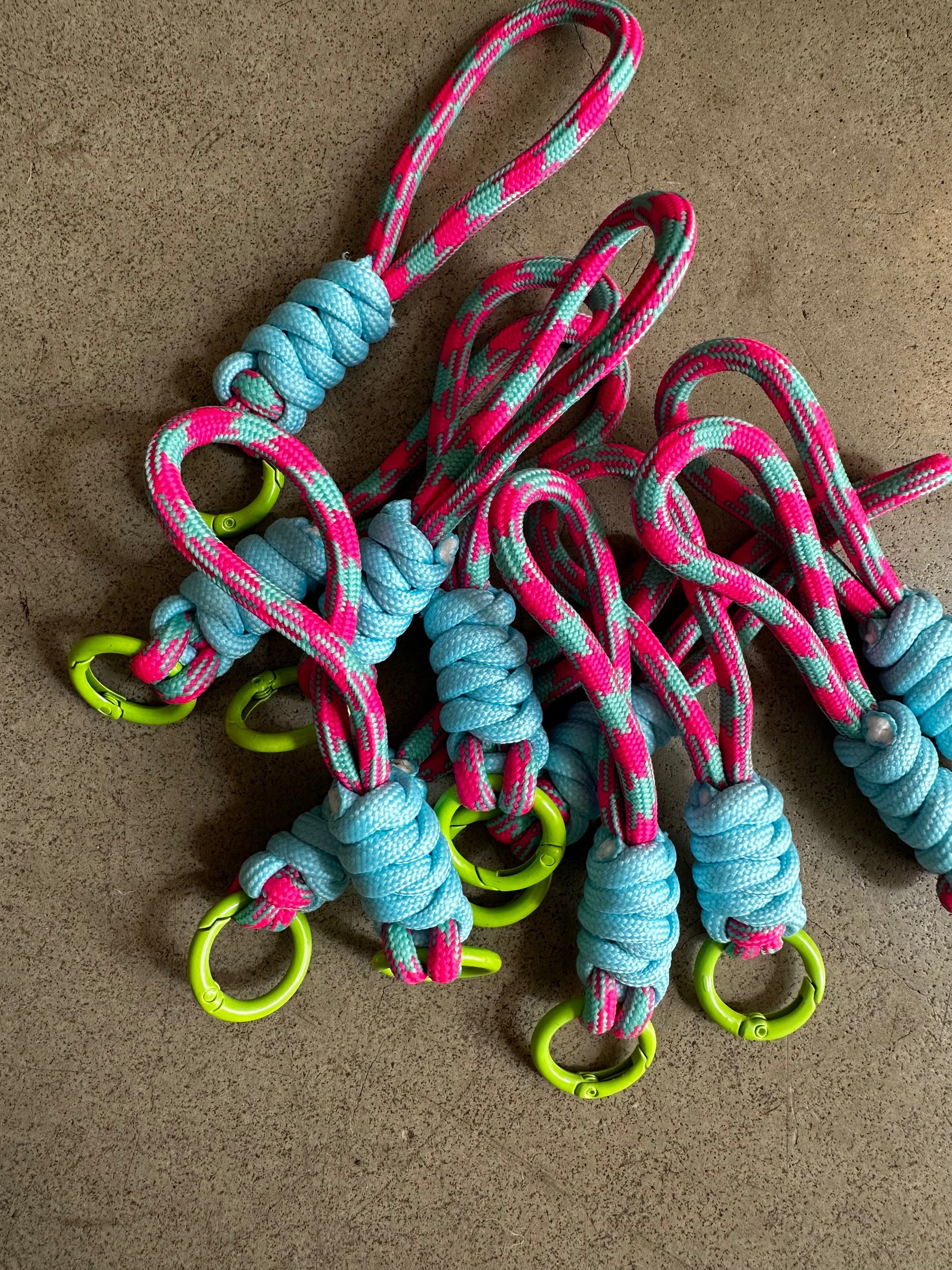 Der LEY! Schlüsselanhänger - blau-pink besteht aus blauen, rosa und grauen geknoteten Kordeln, die an einem leuchtend gelb-grünen Ring befestigt sind - ideal für Taschen oder Schlüssel. Auf einer strukturierten braunen Oberfläche arrangiert, verleiht er Ihren Accessoires einen lebendigen Touch.