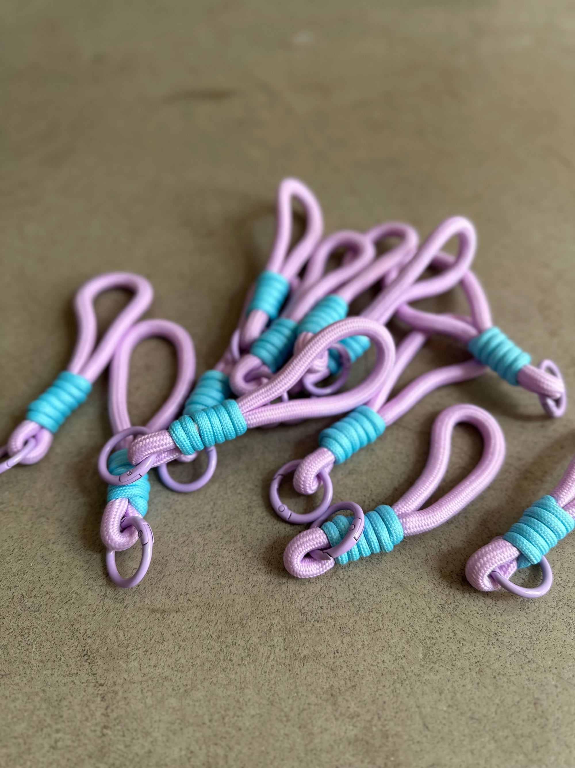 Ein Set aus LEY! Schlüsselanhänger - blau-lila, mit lavendelfarbenen Kordelschlaufen mit leuchtend blauen Wicklungen, liegen auf einer glatten, hellbraunen Oberfläche. Diese handgefertigten Schlüsselanhänger haben ein schlichtes Design, das sie zu einer charmanten Ergänzung für jede Tasche macht.