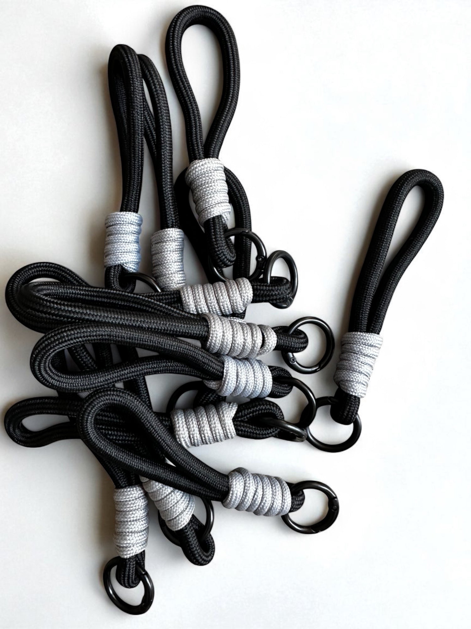 Ein Set von LEY! Schlüsselanhänger - schwarz besteht aus schwarzen Bungee-Kordeln mit Metallringen, grauen und weißen Fäden an den Enden, die sich ideal als Schlüsselanhänger oder Taschenanhänger eignen und auf einer weißen Fläche angeordnet sind.
