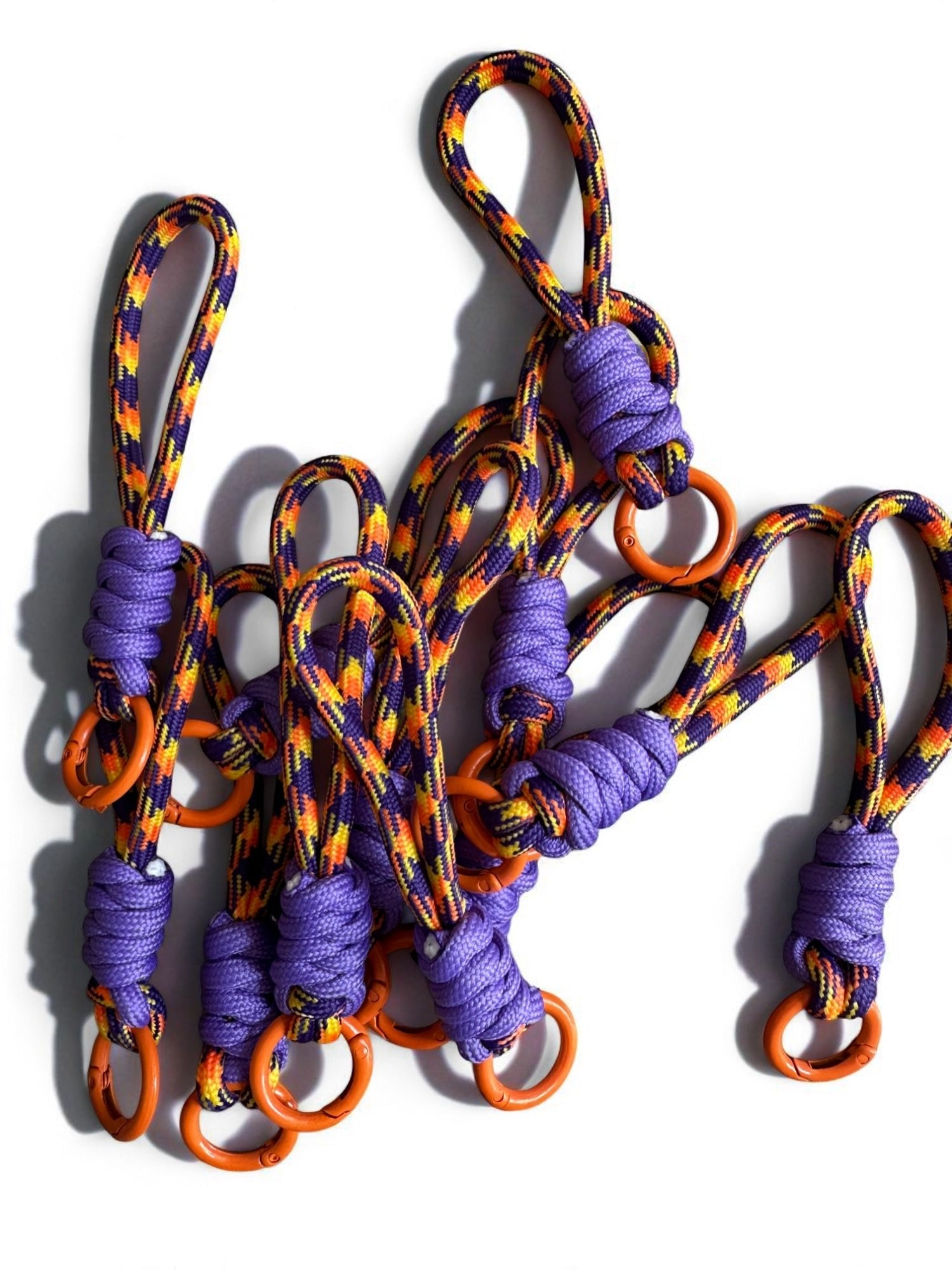 Der LEY! Schlüsselanhänger - lila-orange zeigt bunte Seile in lila, orange, gelb und schwarz mit lila Knoten und orangen Metallringen - ideal für Taschen oder Schlüssel. Arrangiert auf weißem Hintergrund.