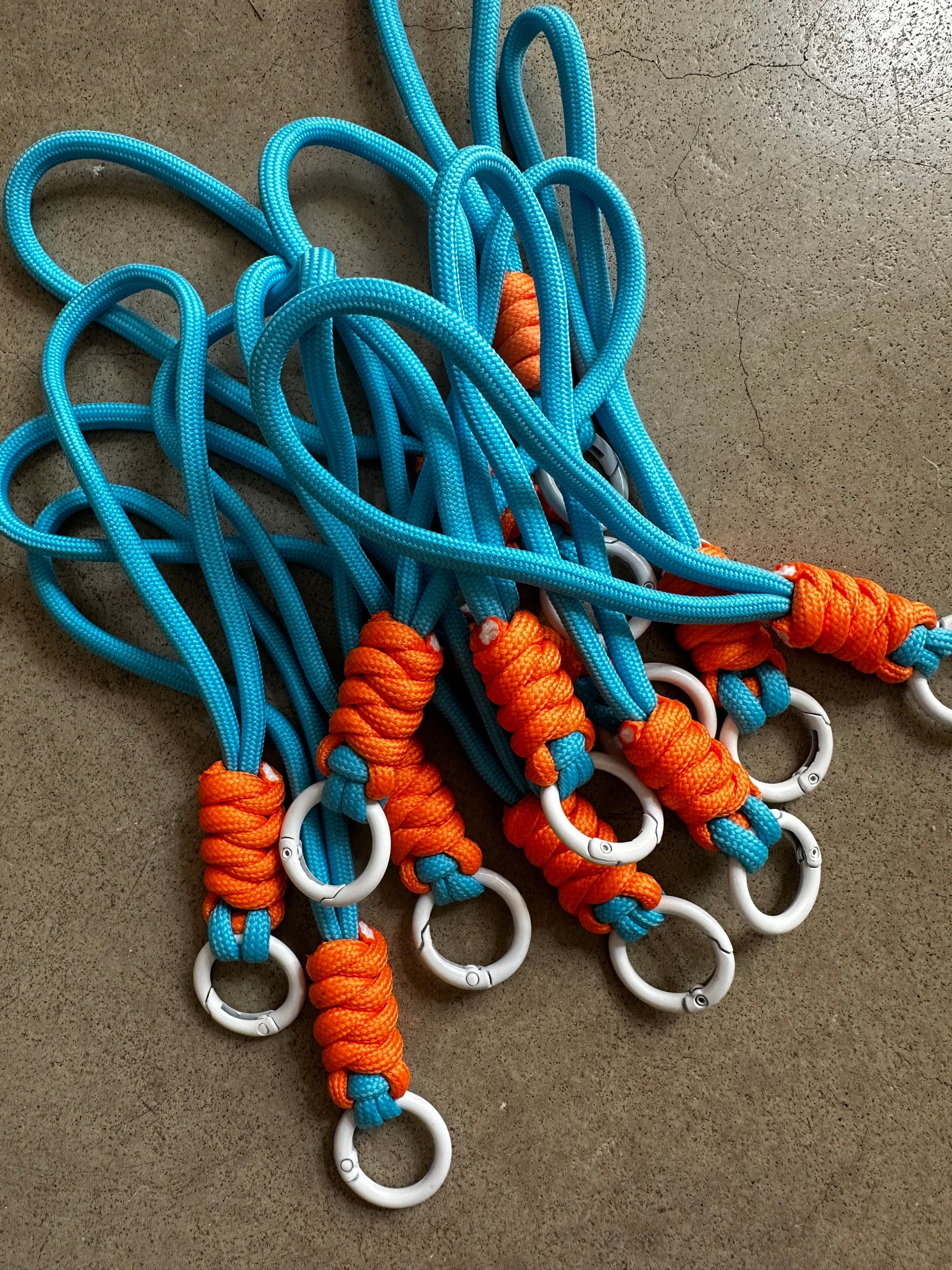 Ein Stapel von LEY! Schlüsselanhänger - orange-blau mit blauen Seilen, orange geknoteten Griffen und weißen Ringen ist auf einer grauen Betonfläche ausgestellt.