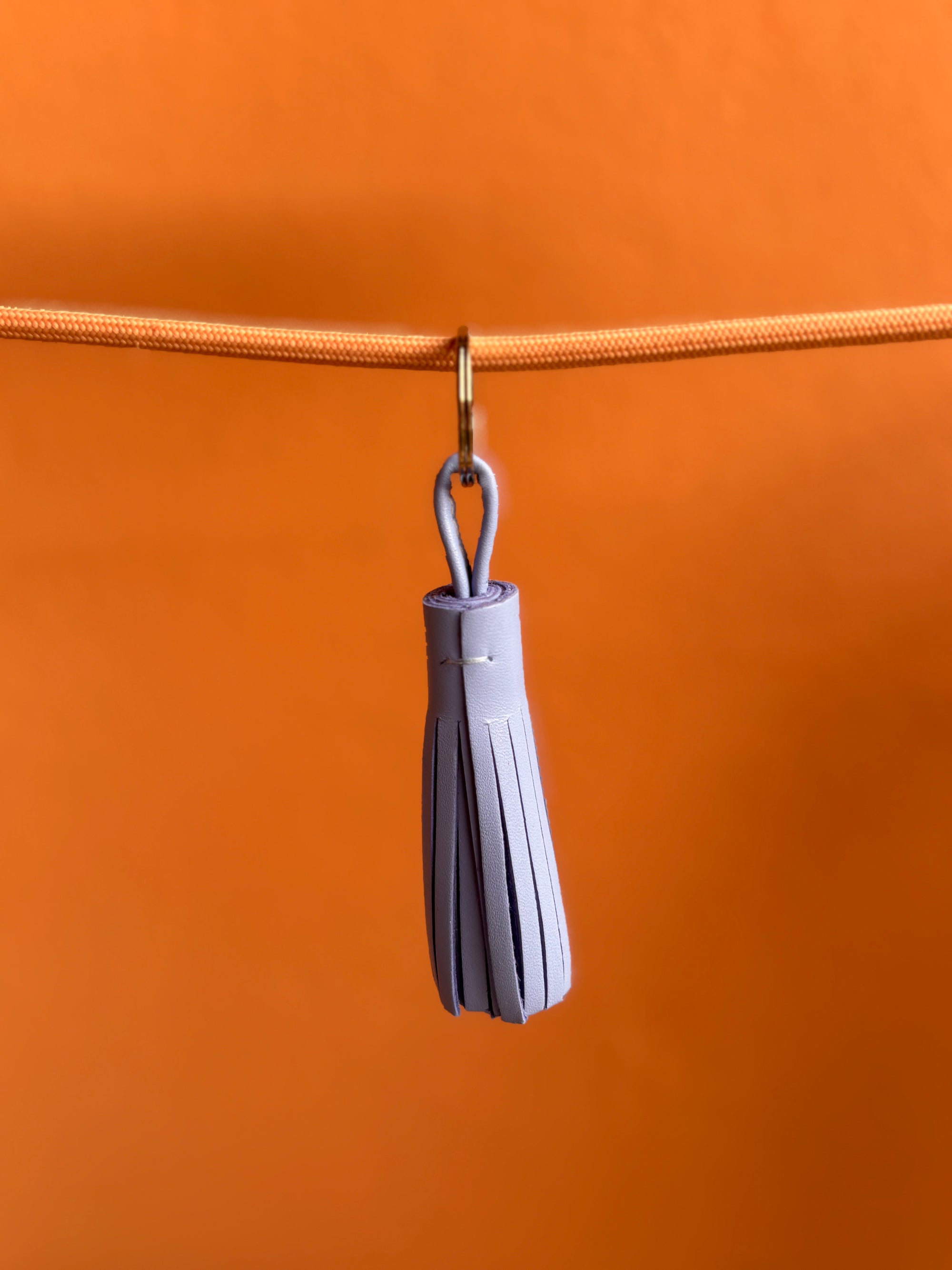 Der Leder-Schlüsselanhänger - lila von LEY! mit helllila Quaste hängt an einem goldenen Ring und orangem Band vor orangefarbenem Hintergrund.