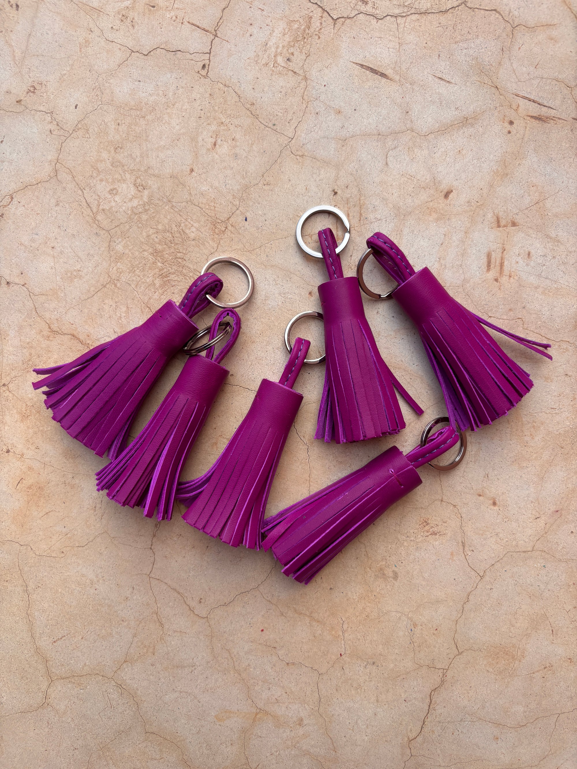 Sechs handgefertigte LEY! Leder-Schlüsselanhänger - magenta mit silbernen Ringen sind auf einer hellbraunen, strukturierten Oberfläche ausgestellt. Ihre gefransten Quasten machen sie zu einer charmanten Geschenkidee für jeden Anlass.