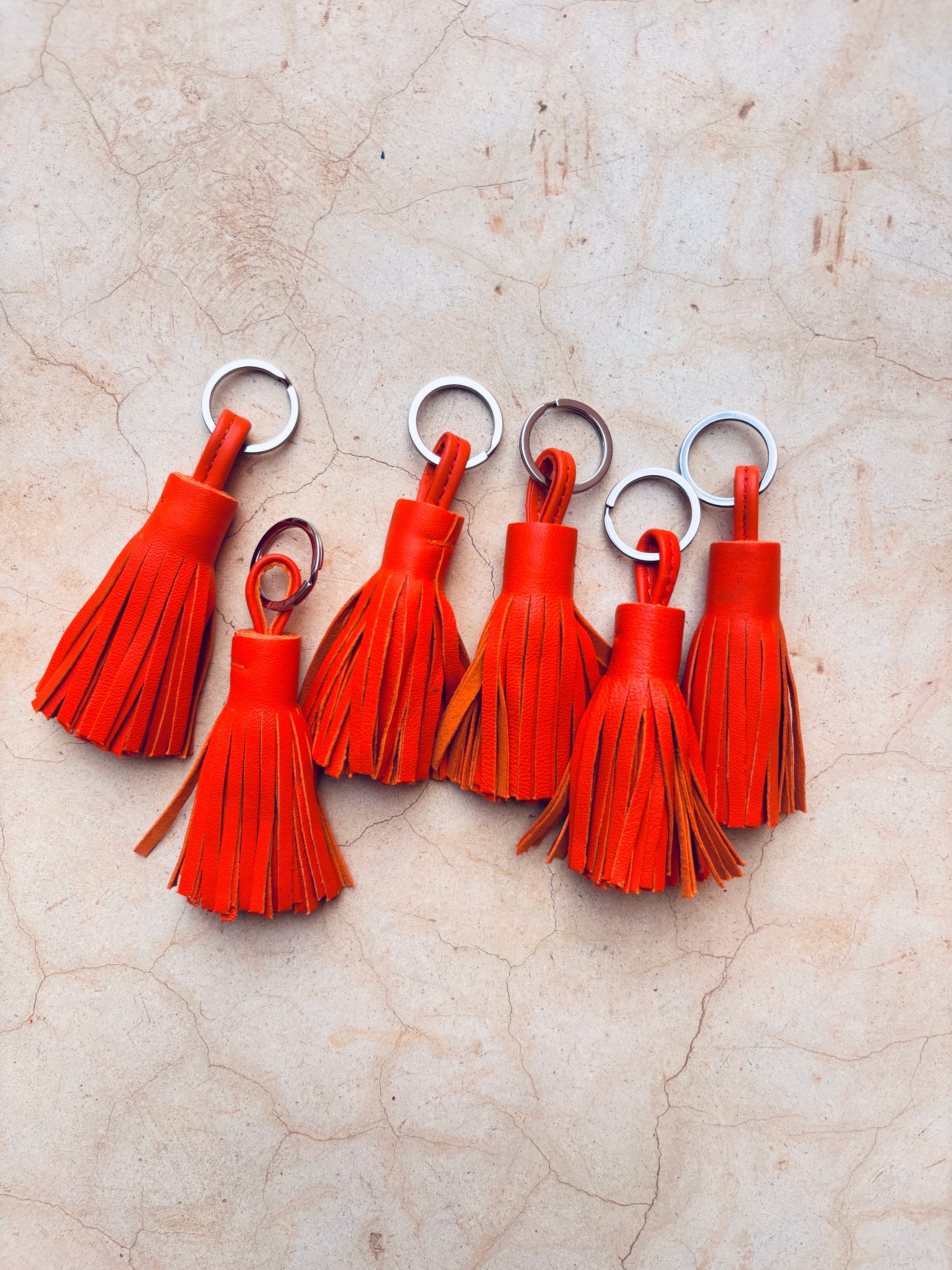 Sechs orange Leder-Schlüsselanhänger von LEY! mit silbernen Ringen liegen auf hellbrauner Oberfläche - eine stilvolle Geschenkidee für jeden Anlass.