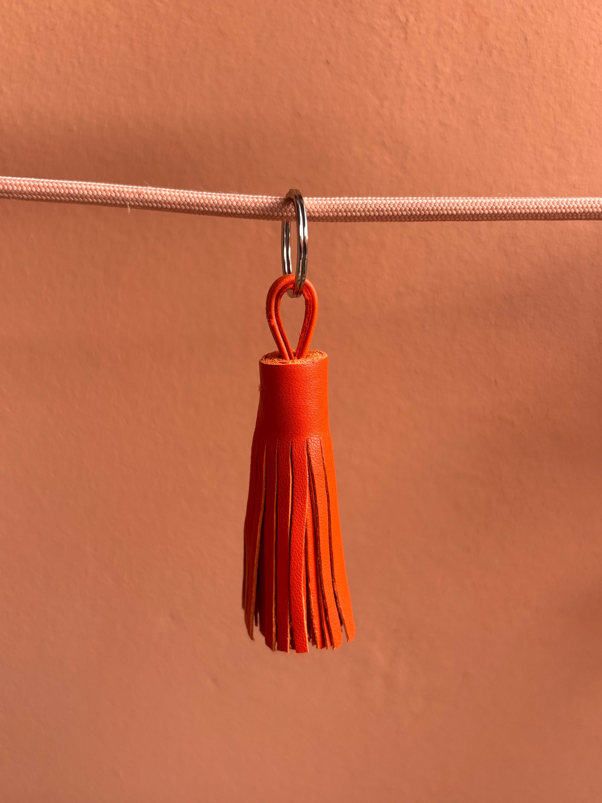 Der Leder-Schlüsselanhänger - orange von LEY! mit Quaste und Schlaufe hängt an einem pinken Band vor einer pfirsichfarbenen Wand - eine stylische Geschenkidee!.