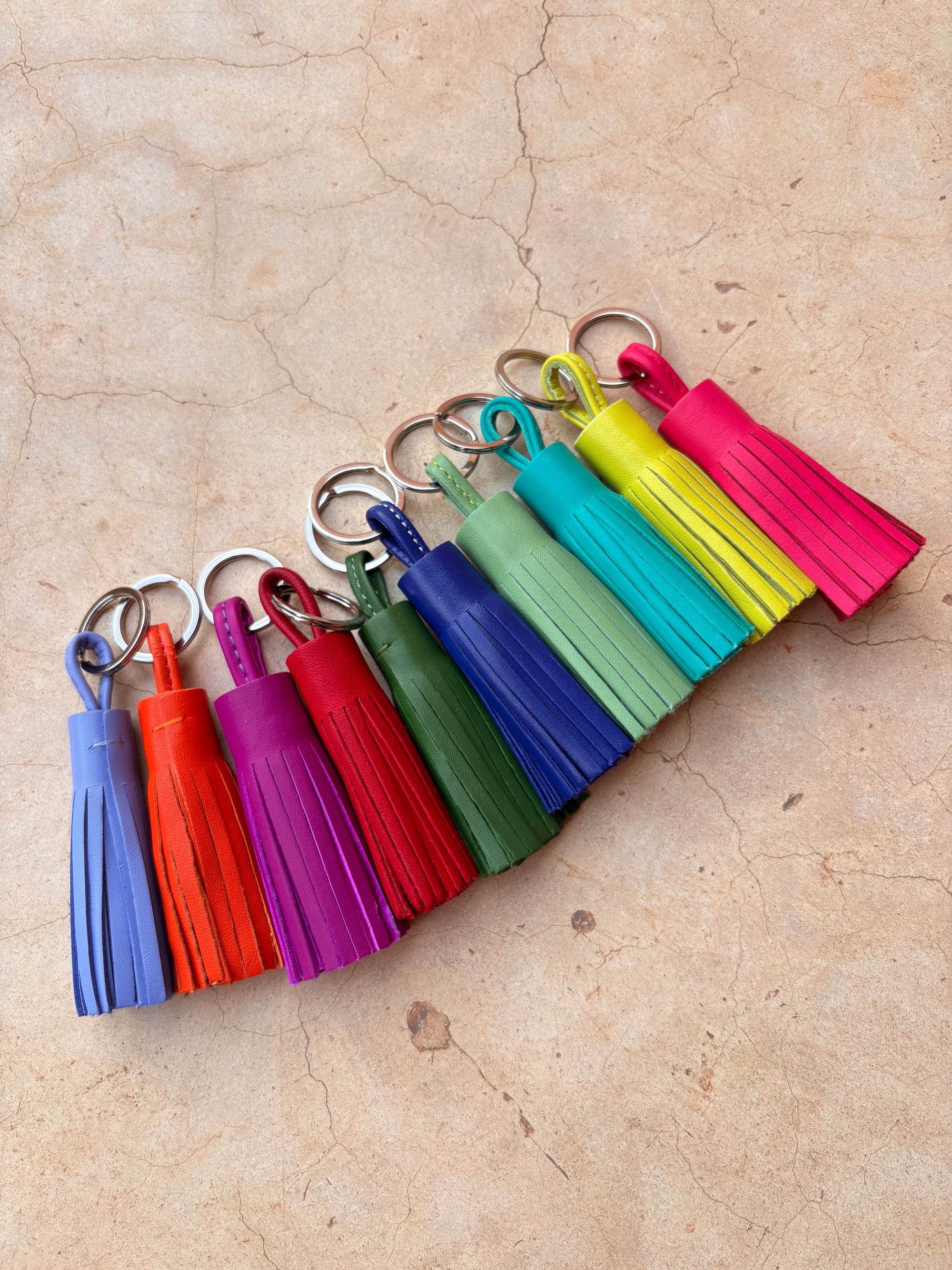 Neun leuchtende LEY! Leder-Schlüsselanhänger - orange, jeder handgefertigt mit einer farbenfrohen Quaste, werden diagonal auf beigefarbenem Rissstein ausgestellt - eine charmante Geschenkidee.