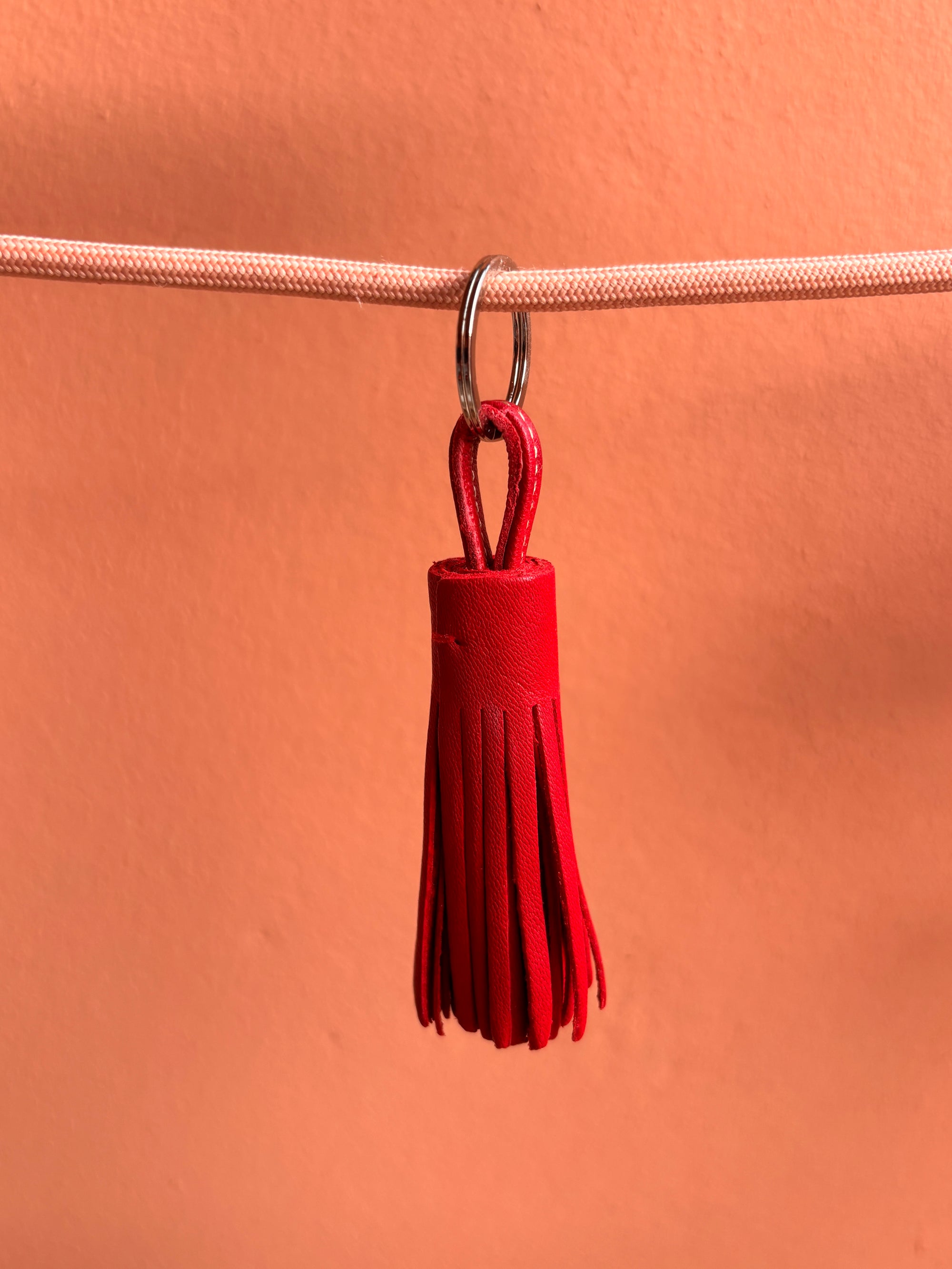 Der Leder-Schlüsselanhänger - rot von LEY! mit roten Lederfransen und Metallring hängt an einem hellen Band - eine stilvolle Geschenkidee vor pfirsichfarbenem Hintergrund.