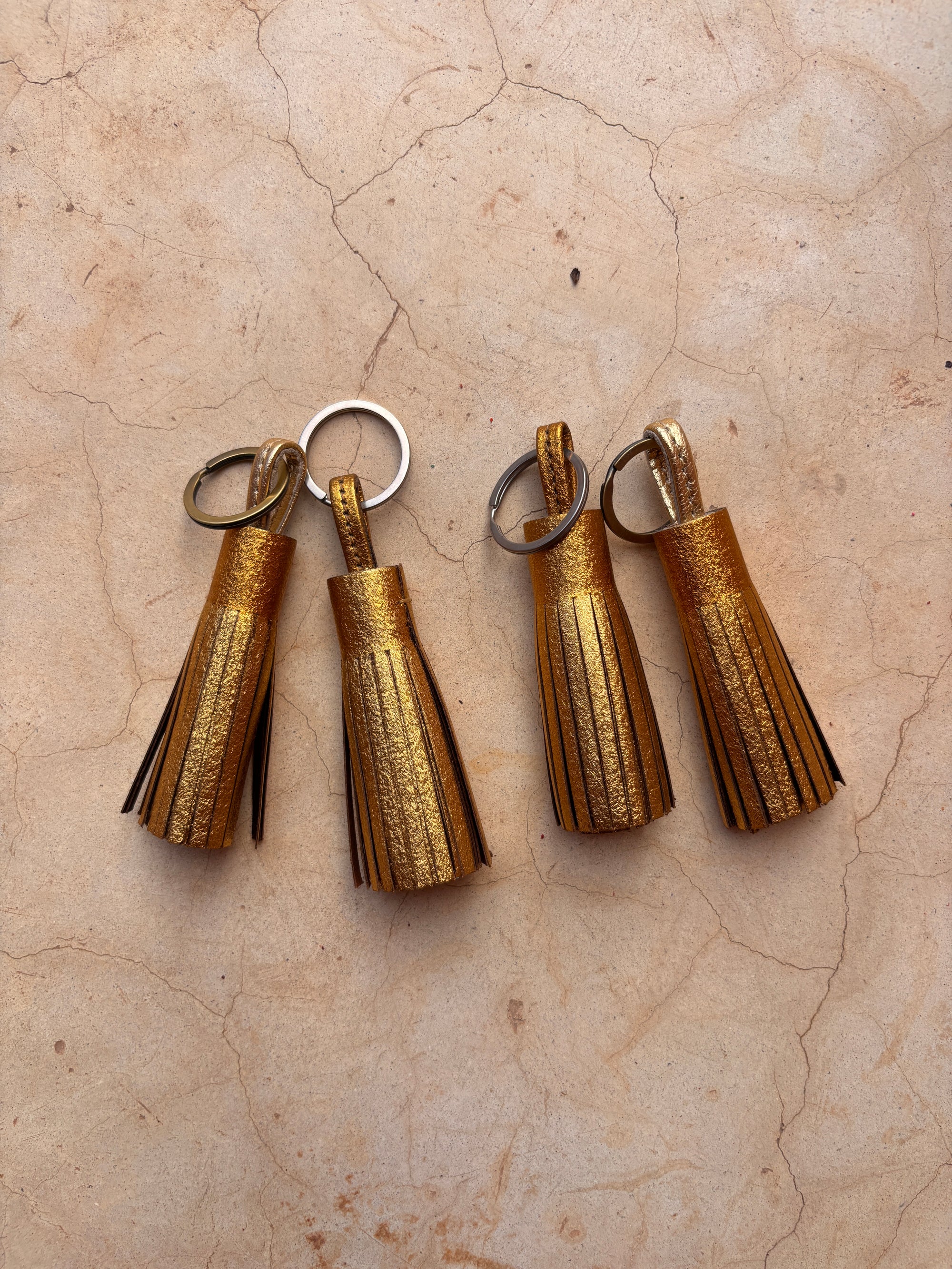 Vier goldfarbene Leder-Schlüsselanhänger - gold by LEY!, jeweils mit einem Metallring, sind auf einer beigen Marmorfläche übersichtlich angeordnet - eine elegante, handgefertigte Geschenkidee für jeden Anlass.
