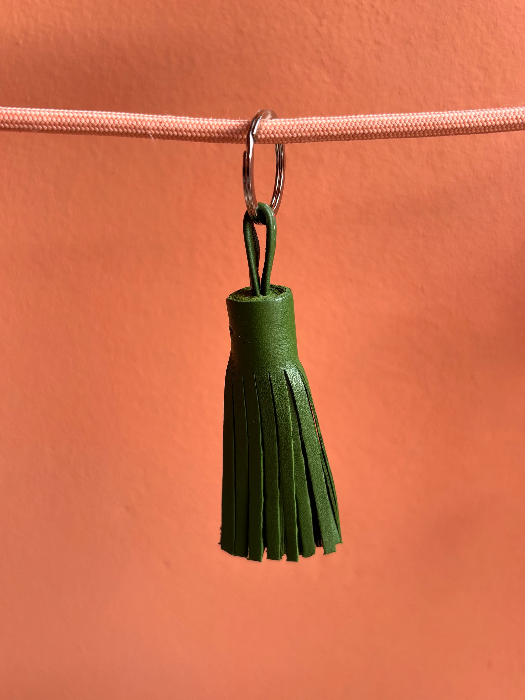 Der LEY! Leder-Schlüsselanhänger - grün ist ein kleines, handgefertigtes Accessoire mit Fransen. An einem pinken Band vor pfirsichfarbenem Hintergrund präsentiert, ist er eine charmante Geschenkidee.