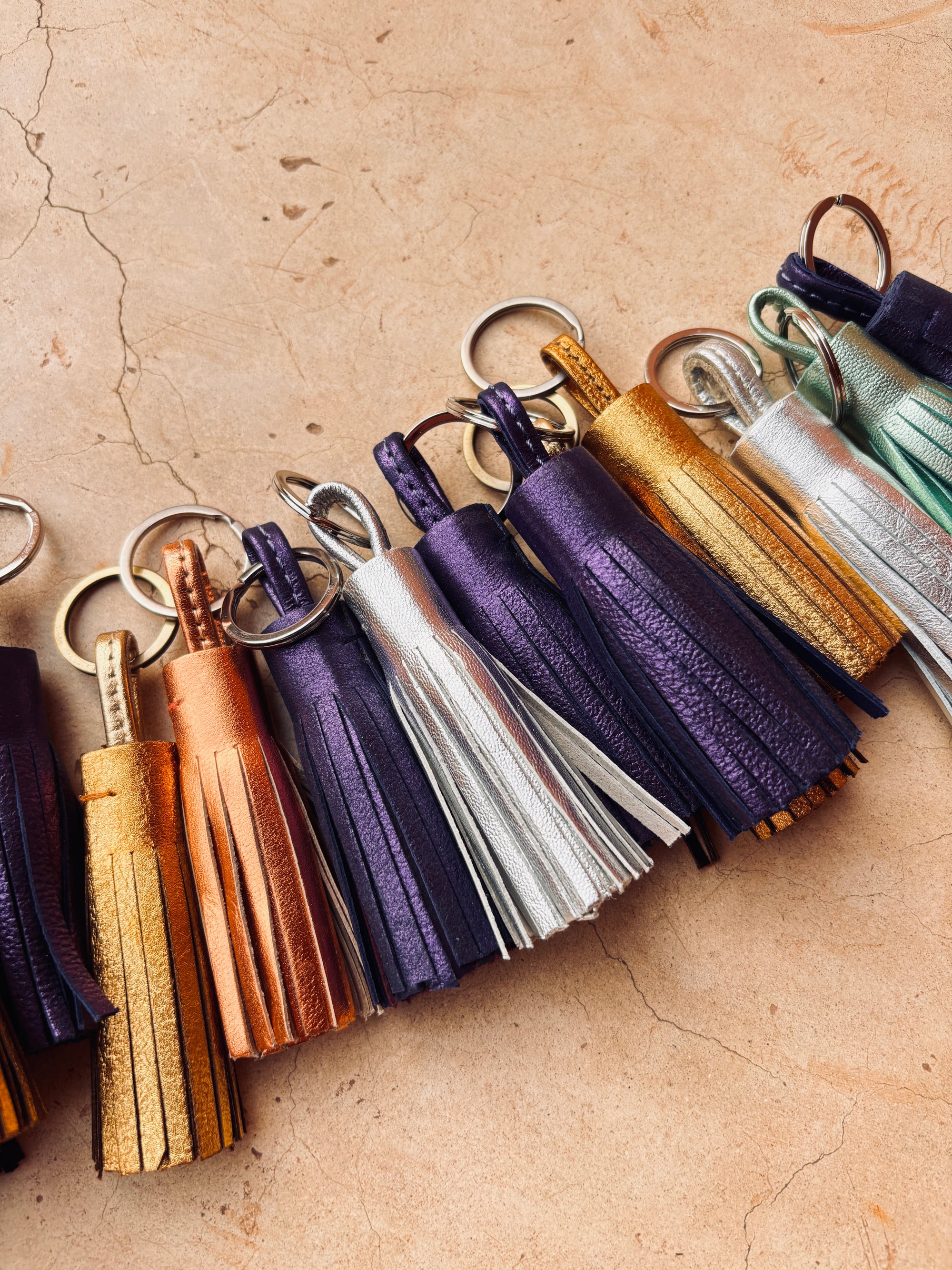 Mehrere LEY! Leder-Schlüsselanhänger - silber mit Quasten in Metallicfarben wie Gold, Lila, Orange, Silber und Türkis liegen diagonal auf einer beigen Oberfläche - eine stilvolle Geschenkidee.