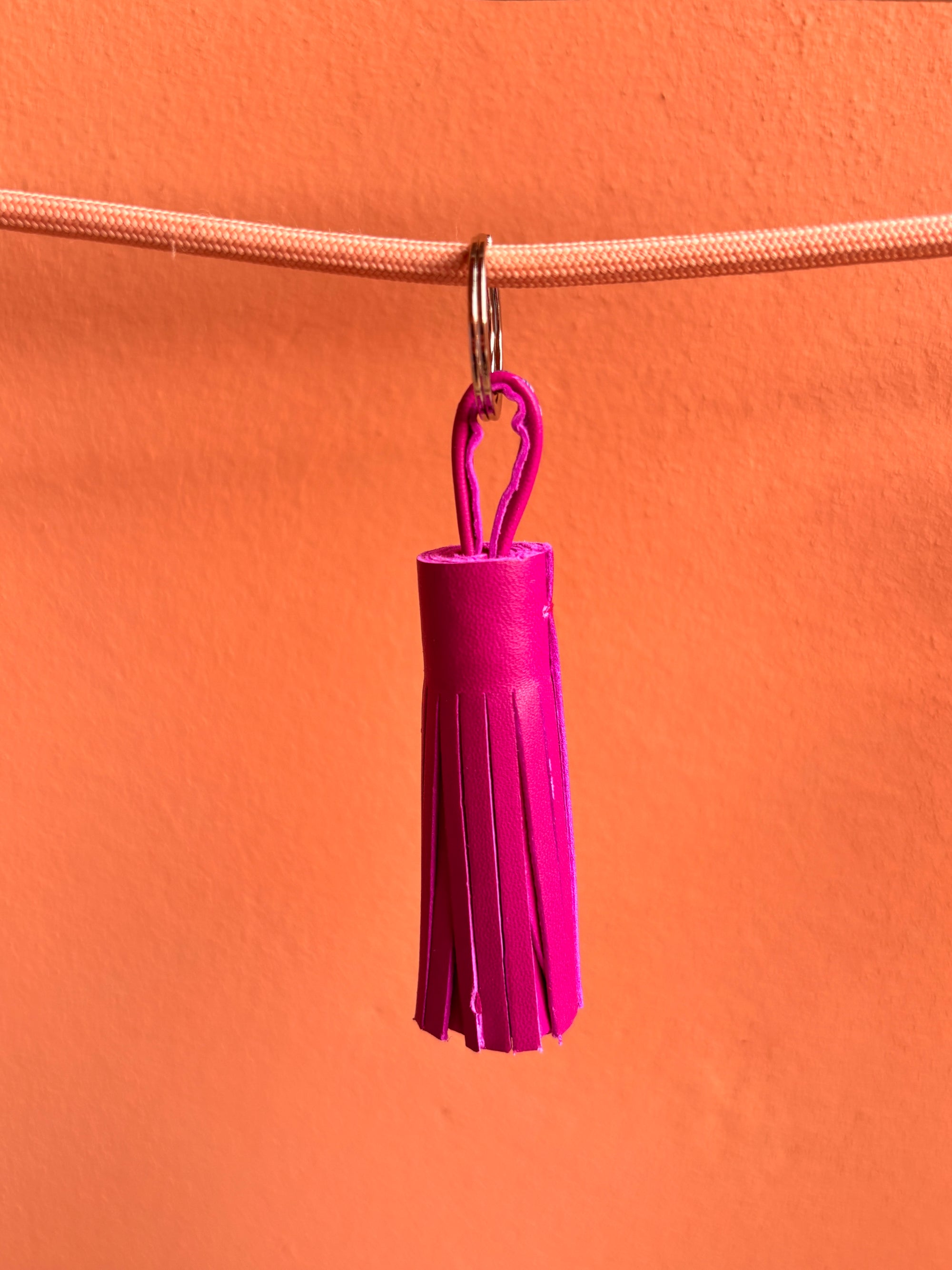 Der LEY! Leder-Schlüsselanhänger - pink mit Quaste hängt an einem goldenen Ring auf pfirsichfarbenem Band vor orangefarbenem Hintergrund - ein stilvolles Geschenk.