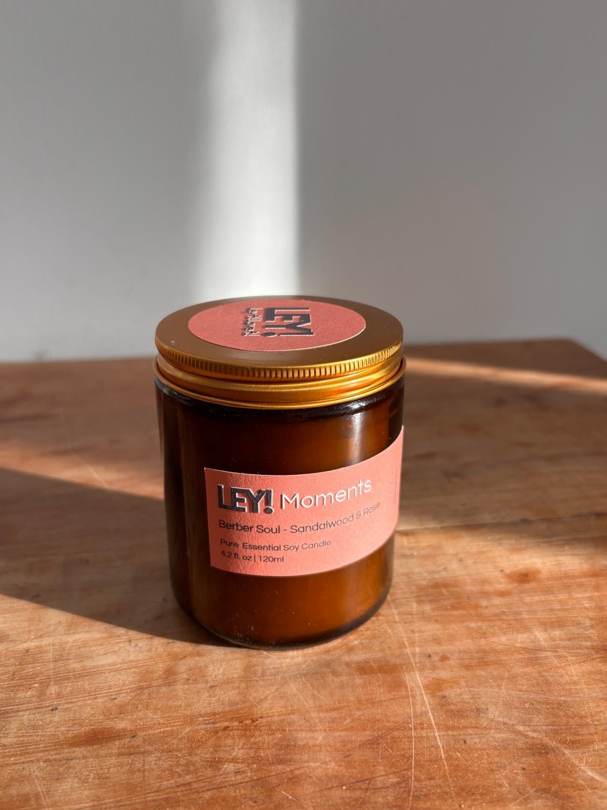 Eine braune Glaskerze mit goldenem Deckel steht auf Holz. Auf dem Etikett steht "LEY! Moments Duftkerze - Marrakesh Souks - Jasmin". Diese LEY! Duftkerze auf Sojabasis leuchtet, wenn das Sonnenlicht sanfte Schatten auf die Oberfläche wirft.