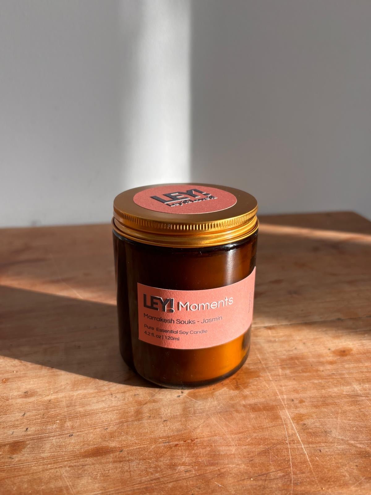 Ein Kerzenglas aus braunem Glas mit goldenem Deckel steht auf Holz, das Sonnenlicht wirft einen Schatten. Auf dem Etikett steht "LEY! Moments Duftkerze - Berber Soul - Sandelholz &amp; Rose". Eine hochwertige LEY! Sojawachskerze für reinen, luxuriösen Duft.