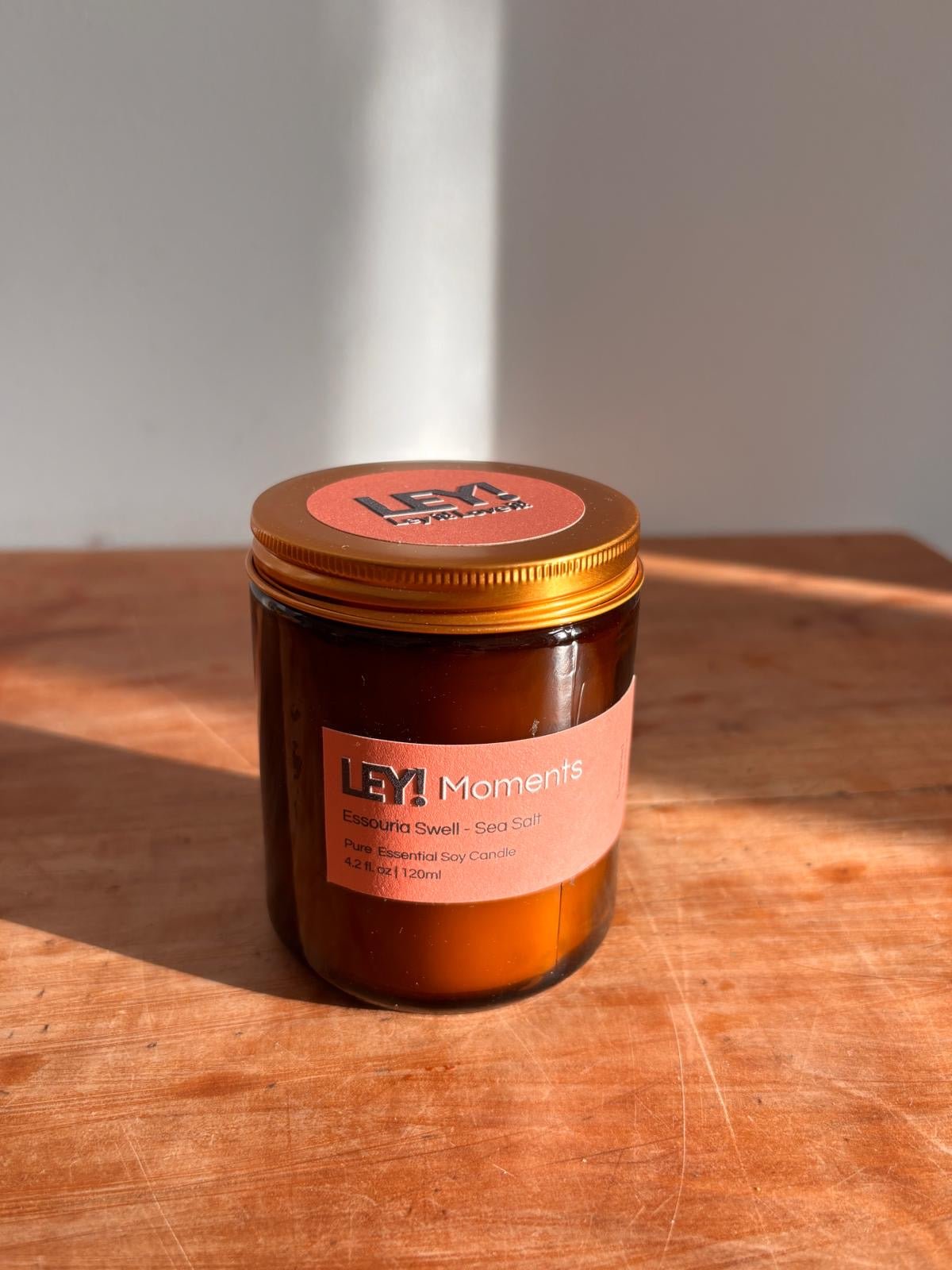 Eine braune Glaskerze mit goldenem Deckel der Marke LEY! steht auf einer Holzfläche im Sonnenlicht. Auf dem Etikett steht "LEY! Moments Duftkerze - Essouria Swell - Sea Salt", was darauf hinweist, dass es sich um eine hochwertige Sojawachskerze handelt.