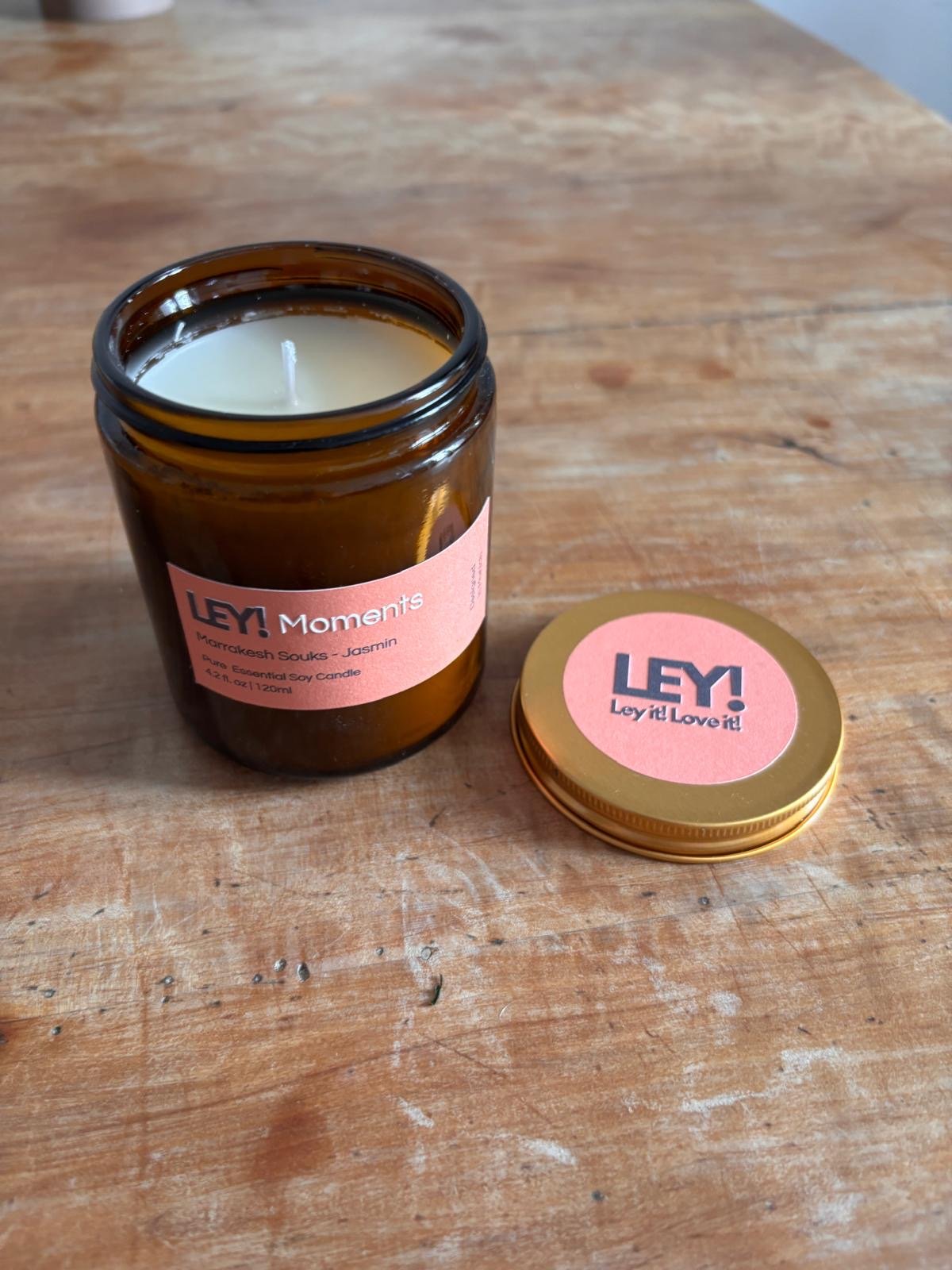 EINE LEY! Moments Duftkerze - Marrakesh Souks - Jasmin von LEY! in einem braunen Glastopf mit pink-orangem Etikett sitzt auf Holz neben seinem goldenen Deckel und hebt elegante, hochwertige sojawachsbasierte Kerzen für einen sauberen Abbrand hervor.