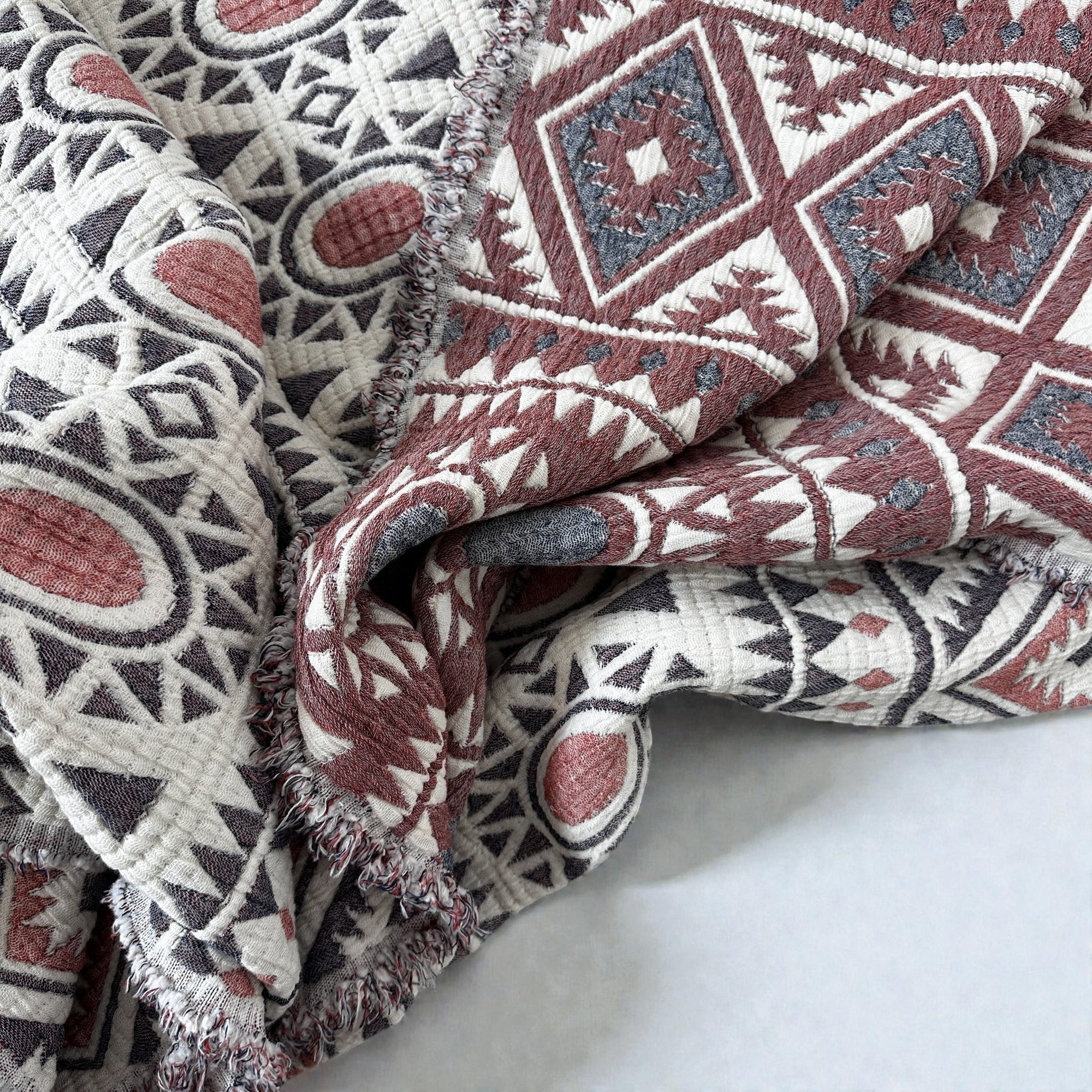Eine Nahaufnahme der LEY! CALMA Musselin Decke (2,30 x 2,60 m) in Rot-Grau-Weiß zeigt ihr Boho-Design mit geometrischen Mustern. Der atmungsaktive, leicht zerknitterte Stoff unterstreicht seine Qualität und seinen Stil.