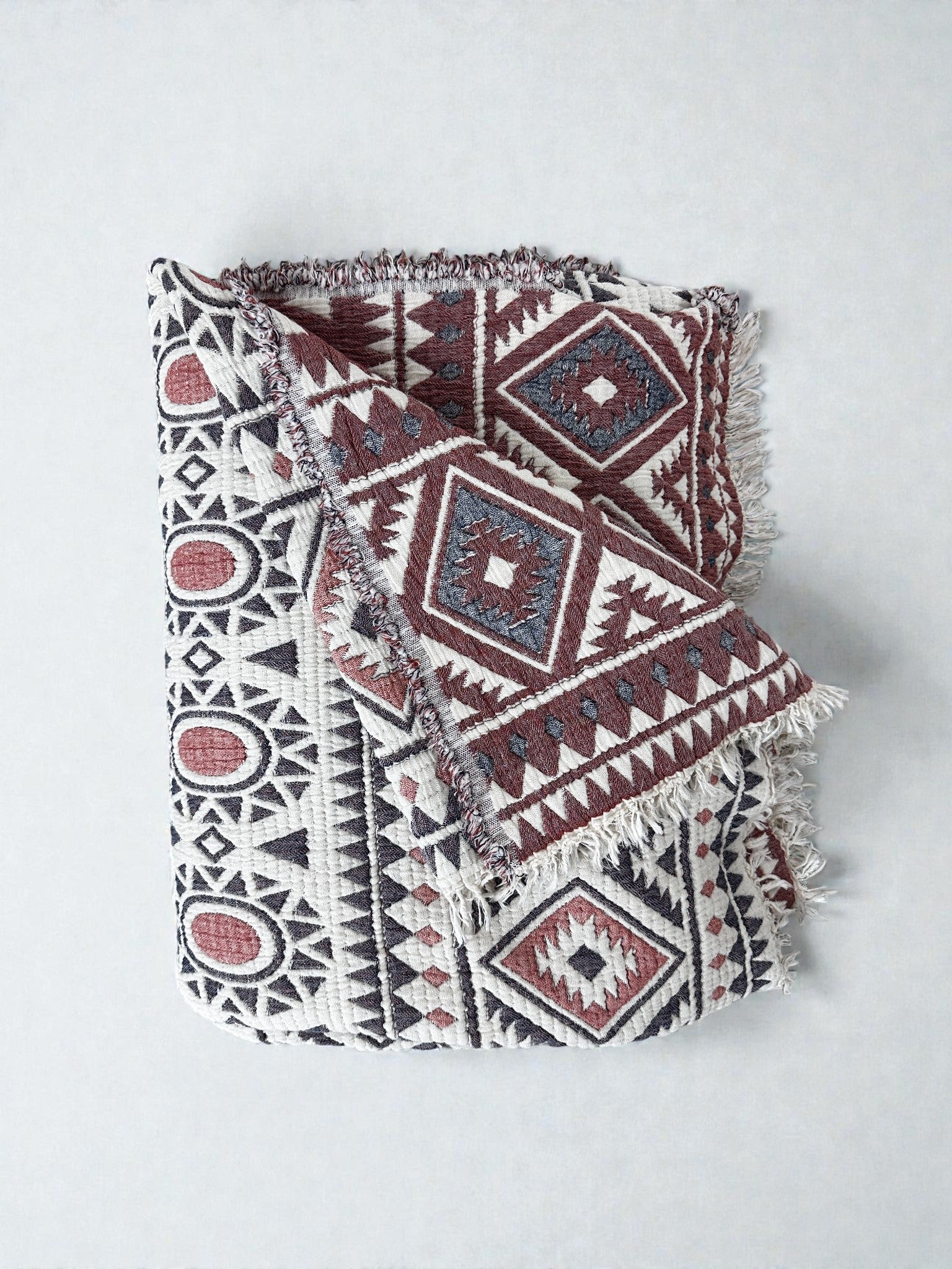 Die CALMA Musselin Decke von LEY! in den Farben Rot, Grau und Weiß (2,30 x 2,60 m) zeigt ein vom Boho-Stil inspiriertes geometrisches Muster mit Rauten, Dreiecken, Kreismotiven und Fransen, das gefaltet auf einem hellen Untergrund liegt.