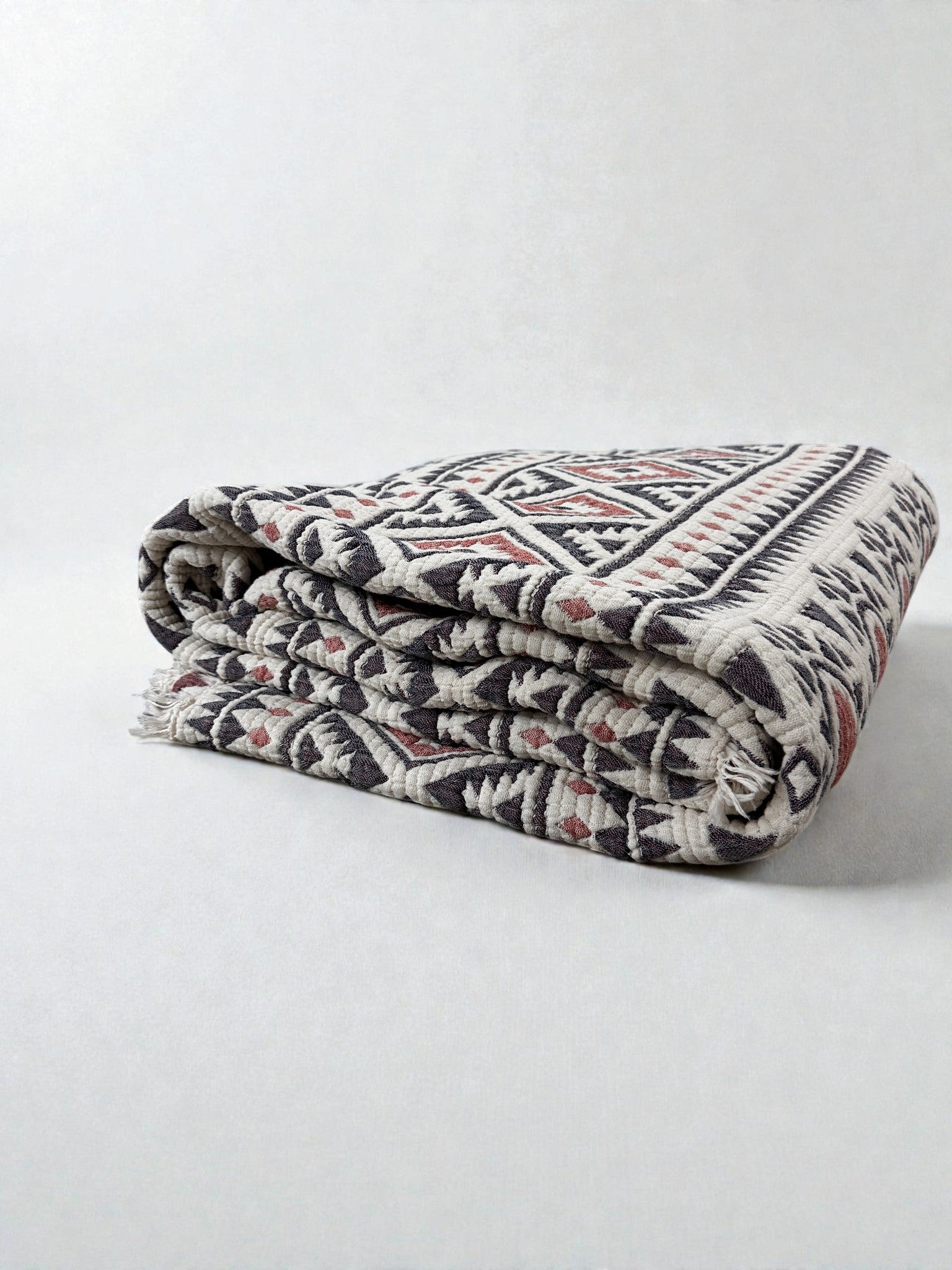 Die CALMA Musselin Decke von LEY! (2,30 x 2,60 m) in Rot-Grau-Weiß hat ein geometrisches Boho-Design und ist weich, stilvoll und atmungsaktiv für ganzjährigen Komfort. Abgebildet gefaltet auf einem unifarbenen, hellen Untergrund.