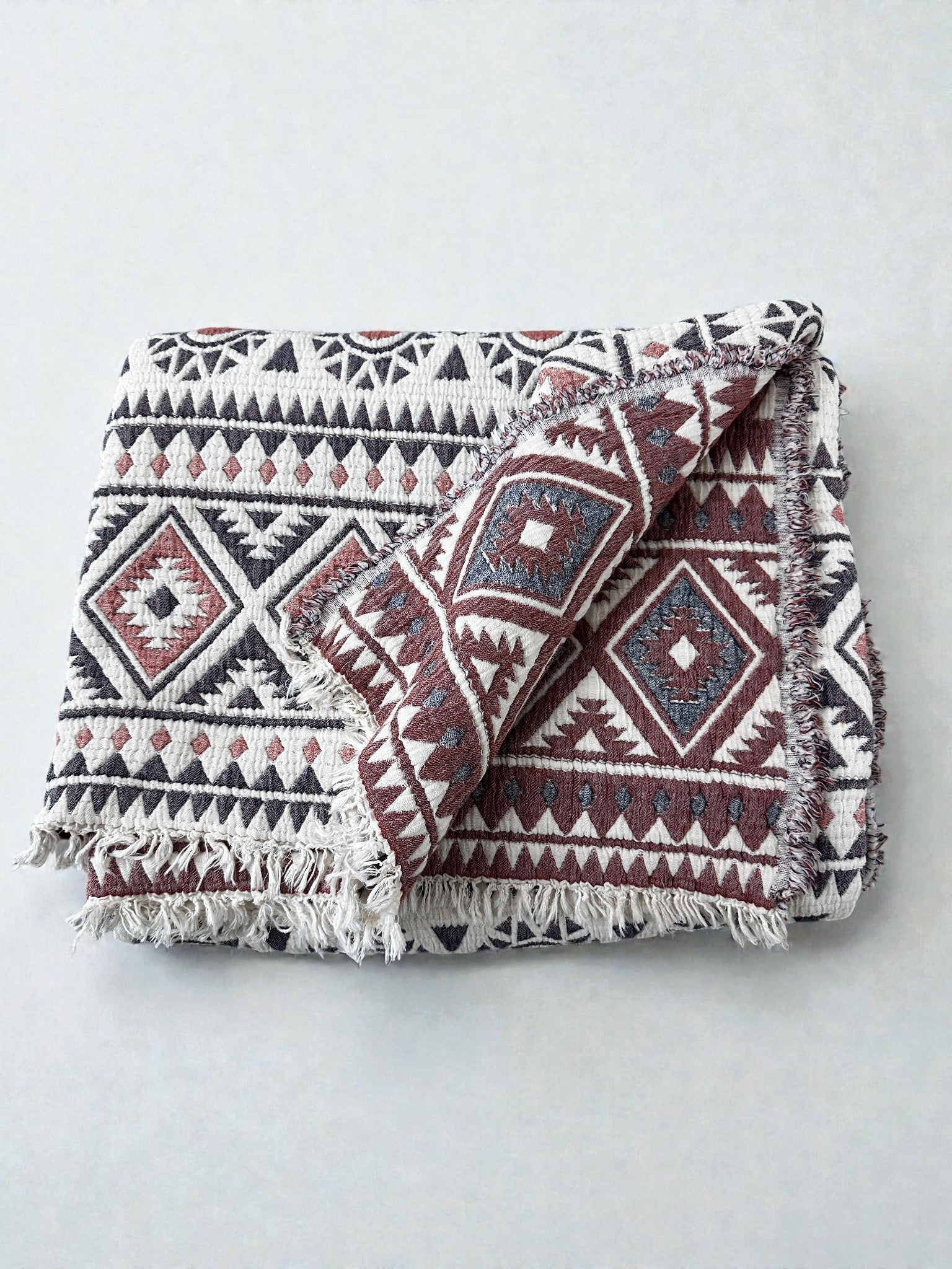 Die CALMA Musselin Decke von LEY! (2,30 x 2,60 m) in Rot-Grau-Weiß zeigt ein geometrisches Tribal-Muster mit Fransenkanten und Boho-Design, sauber gefaltet auf einem hellen neutralen Hintergrund.