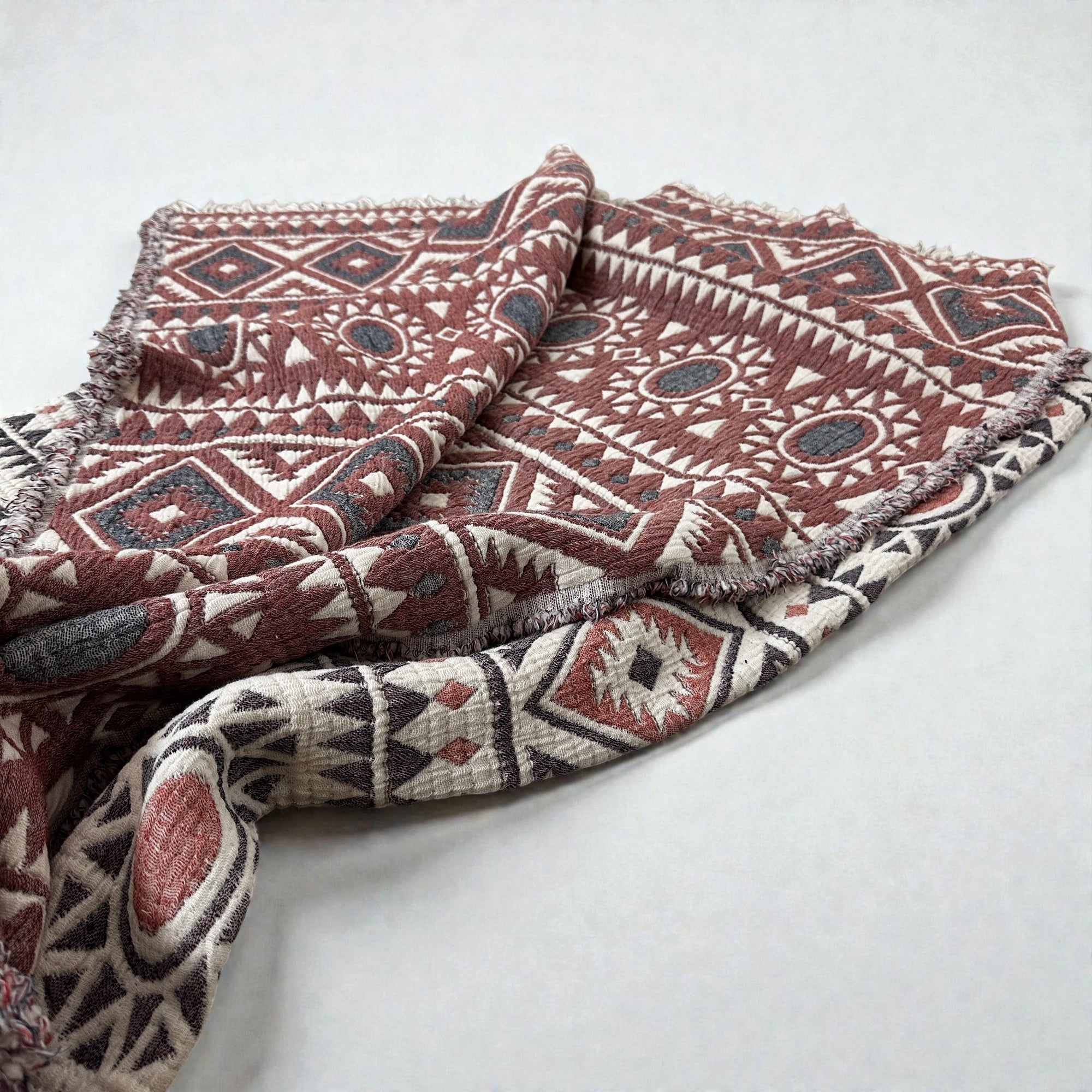 Die CALMA Musselin Decke von LEY! (2,30 x 2,60 m) in Rot-Grau-Weiß zeigt geometrische Muster im Boho-Design - Diamanten, Dreiecke und Kreise - auf neutralem Hintergrund; stilvoll und atmungsaktiv für jeden Raum.