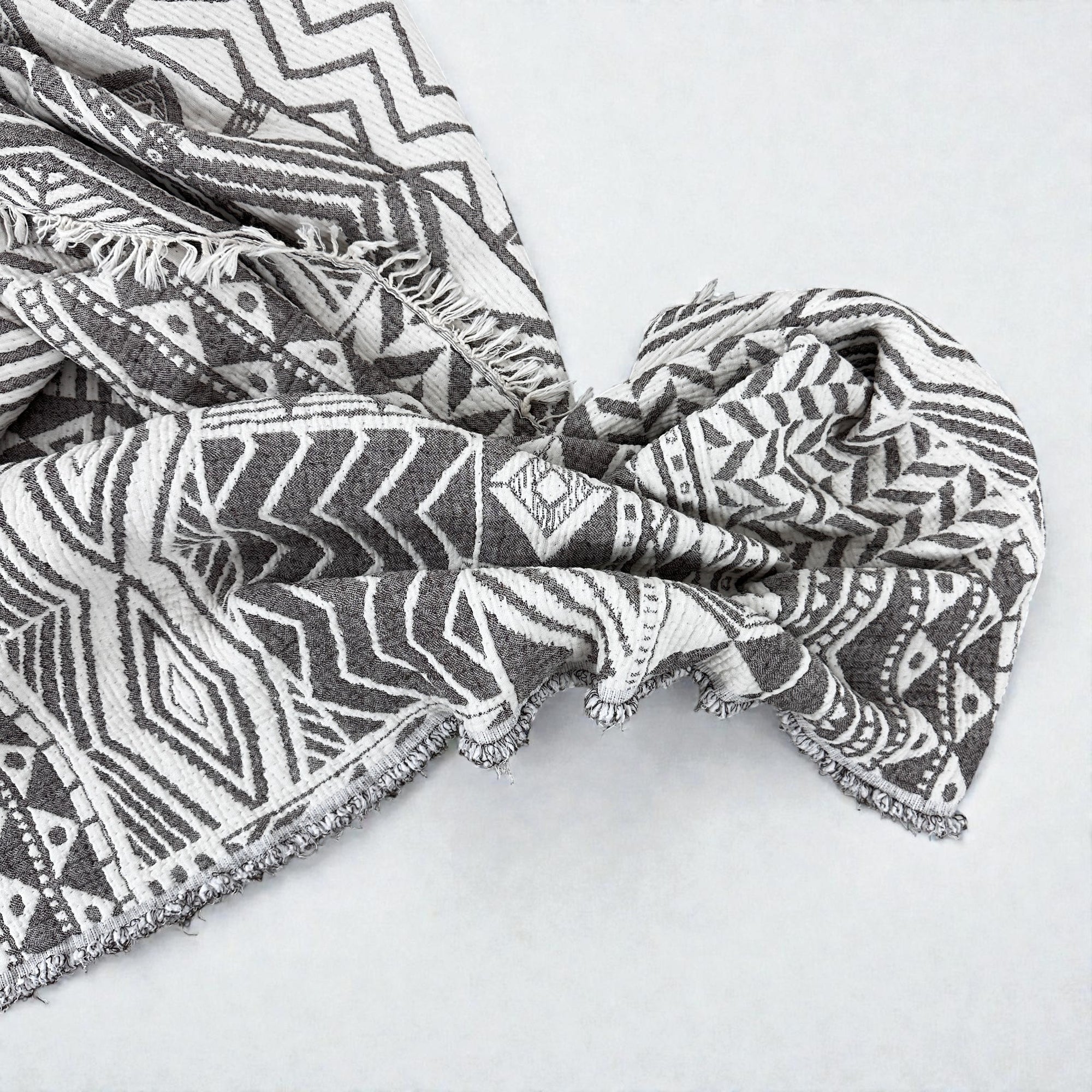 Die CALMA Musselin Decke von LEY! (1,50 x 2,40 m) in Graubraun-Weiß begeistert mit weich fließendem Stoff, Boho-Design, schwarzen und weißen geometrischen Mustern sowie Fransen - für einen trendigen Look im modernen Zuhause.