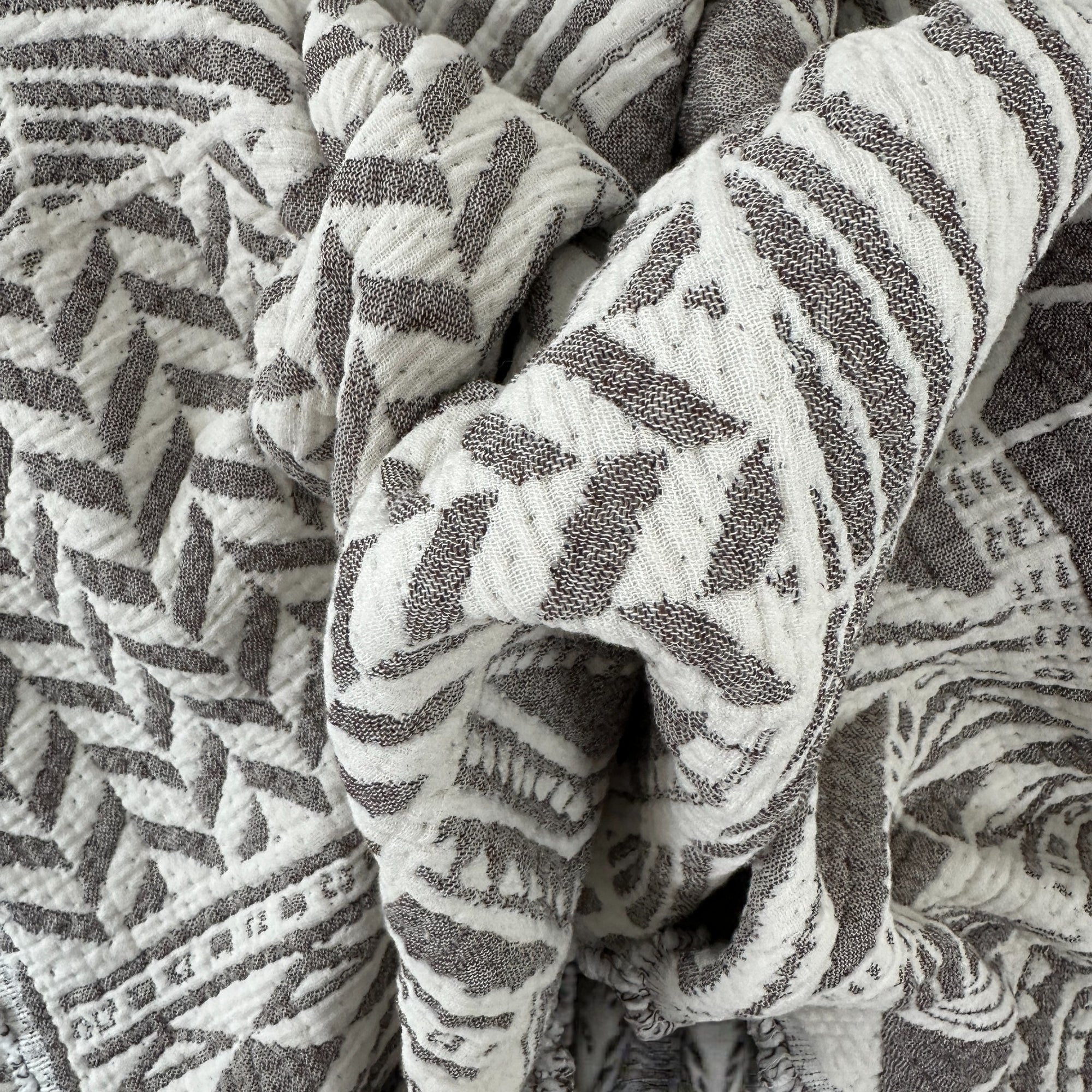 Eine Nahaufnahme der CALMA Musselin Decke von LEY! in Graubraun-Weiß (1,50 x 2,40 m) zeigt ihre weiche, strukturierte Oberfläche mit abstraktem geometrischen und zickzackförmigen Boho-Design für einen haptischen Reiz.