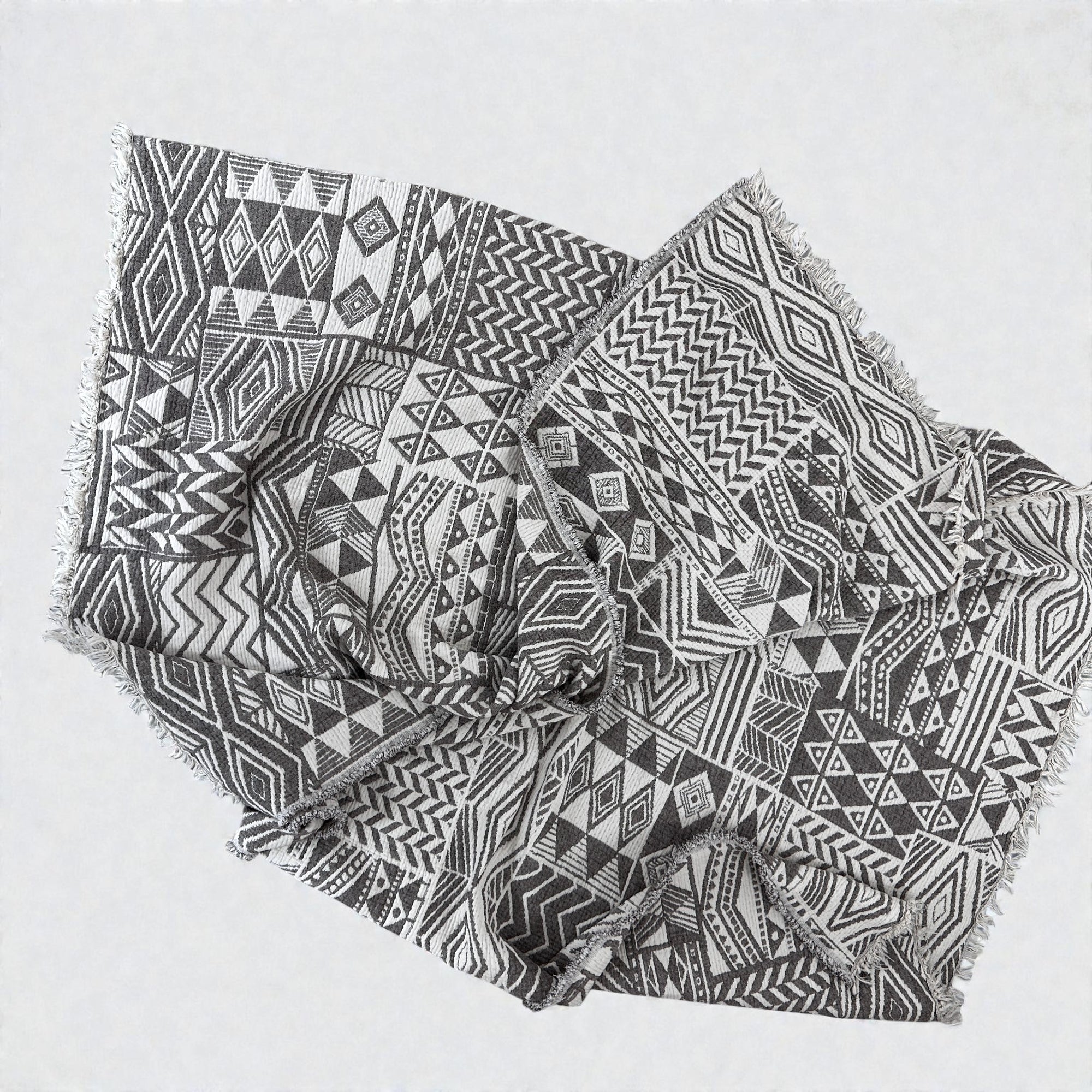 Die CALMA Musselin Decke von LEY! in graubraun-weiß (1,50 x 2,40 m) hat ein aufwendiges geometrisches Boho-Design mit ausgefransten Kanten und fühlt sich superweich an, wenn sie locker gefaltet auf hellem Grund liegt.