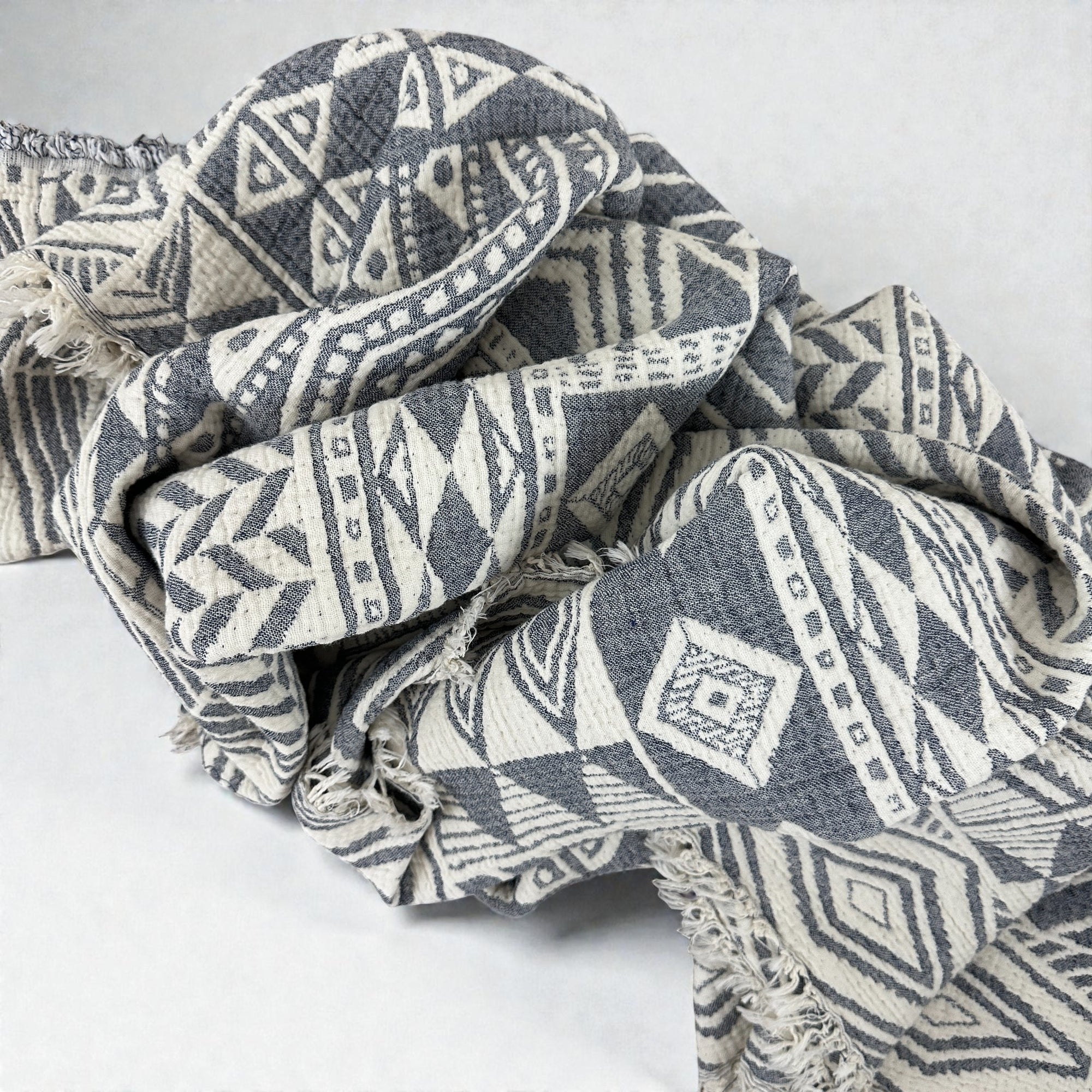 Die CALMA Musselin Decke von LEY! in Grau-Weiß (1,50 x 2,40 m) zeigt ein geometrisches Rauten- und Dreiecksmuster mit ausgefransten Kanten und Boho-Vibes, das sich zerknittert vor einem unifarbenen, hellen Hintergrund abzeichnet.