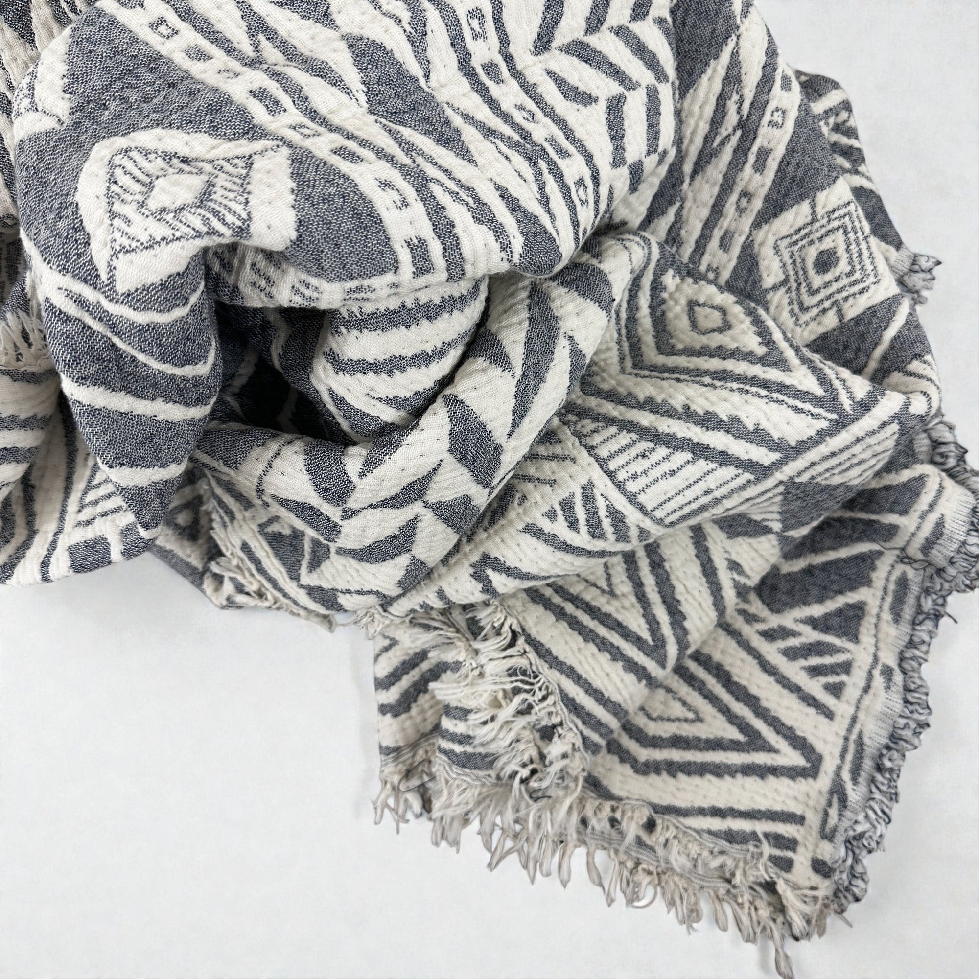 Eine Nahaufnahme der LEY! CALMA Musselin Decke in Grau-Weiß (1,50 x 2,40 m) zeigt ein weiches, atmungsaktives Gewebe mit filigranen geometrischen Mustern und fransigen Kanten - perfekt für Boho-Designs.