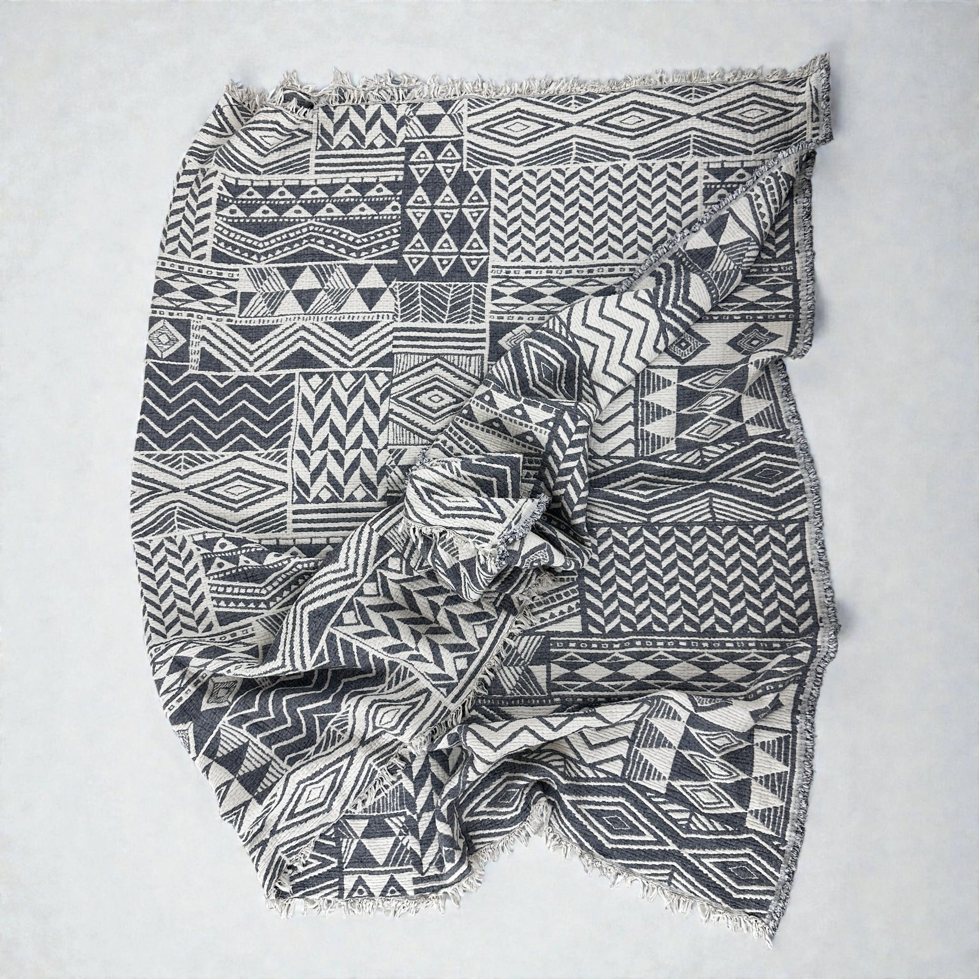 Die CALMA Musselin Decke von LEY! in Grau-Weiß (1,50 x 2,40 m) besticht durch schwarz-weiße geometrische Muster, leicht zerknitterte Fransenkanten und ein atmungsaktives Boho-Design, das auf einer ebenen Fläche ausgebreitet wird.
