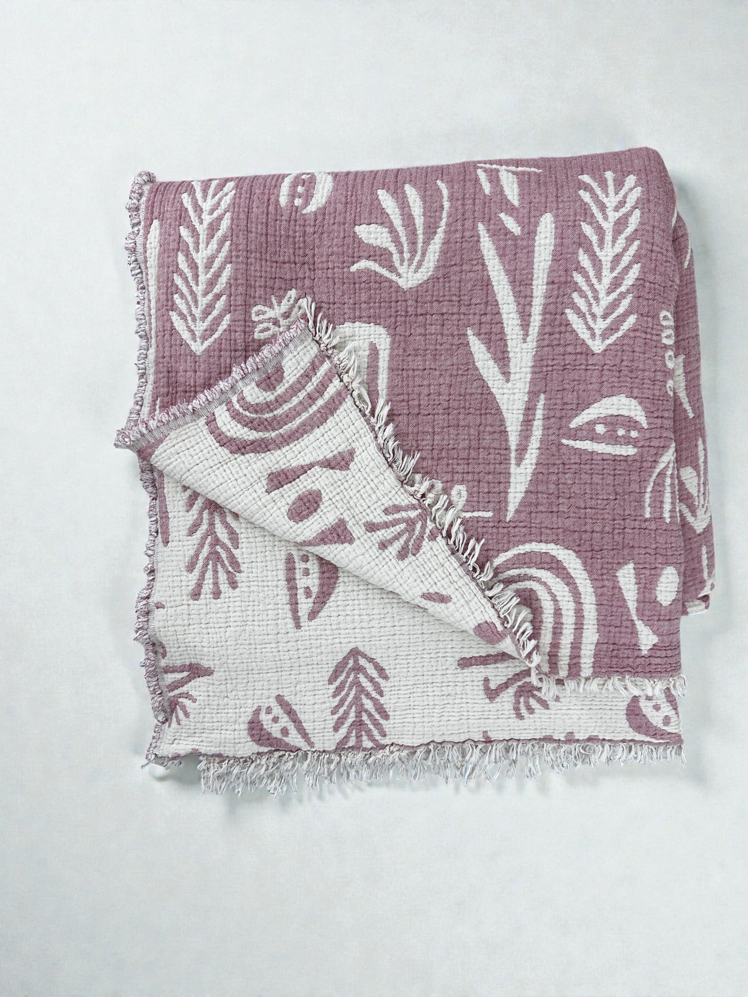 Die CALMA Musselin Decke von LEY! in Rosé-Weiß (2,30 x 2,40 m) zeichnet sich durch eine strukturierte, atmungsaktive Oberfläche mit abstrakten Pflanzen- und Regenbogenmotiven, Boho-Design-Akzenten, Fransenrändern und einem wendbaren Design aus.