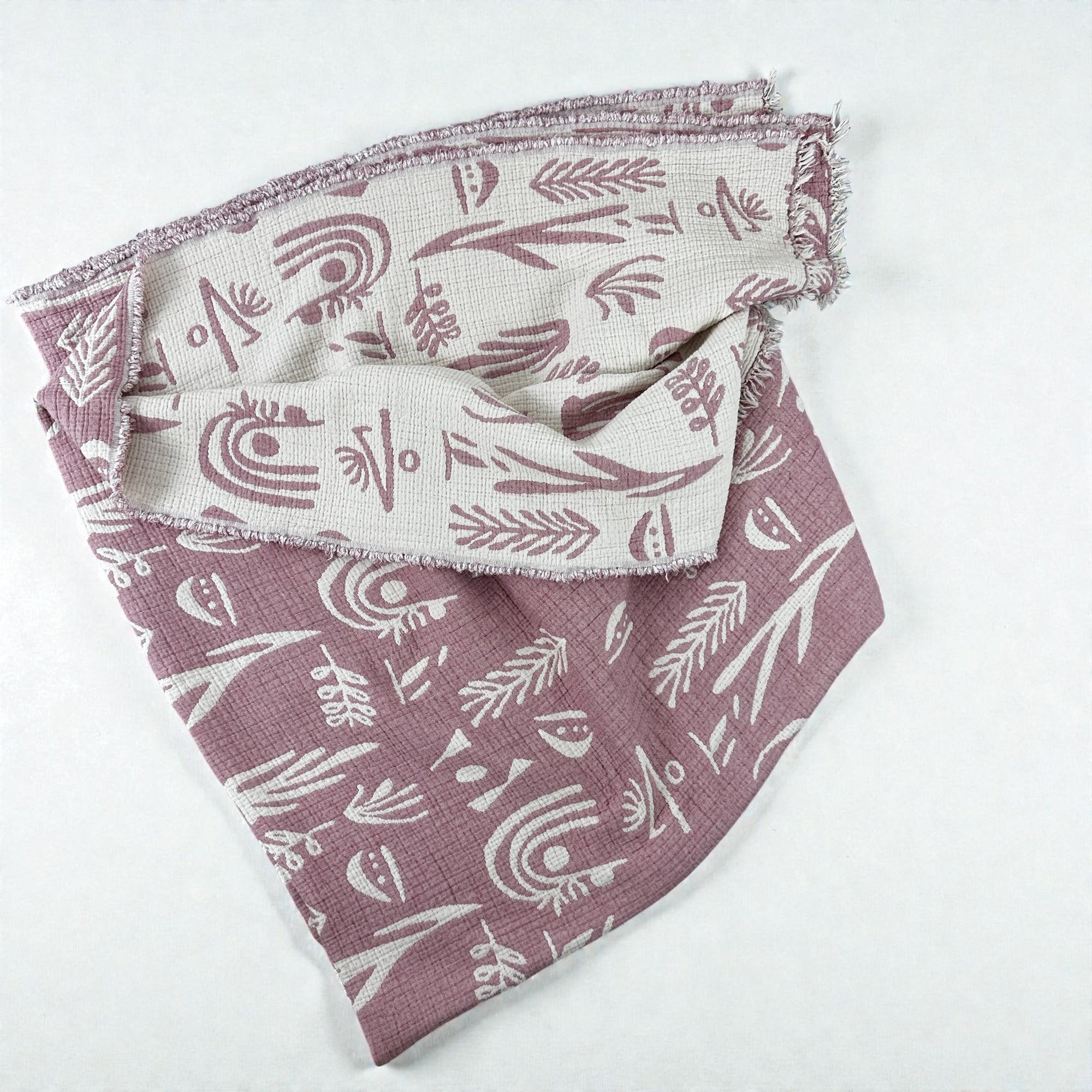 Die LEY! CALMA Musselin Decke in Rosé-Weiß (2,30 x 2,40 m) zeigt ein abstraktes Boho-Muster mit ausgefransten Kanten und ist auf einer leichten Oberfläche locker gefaltet.
