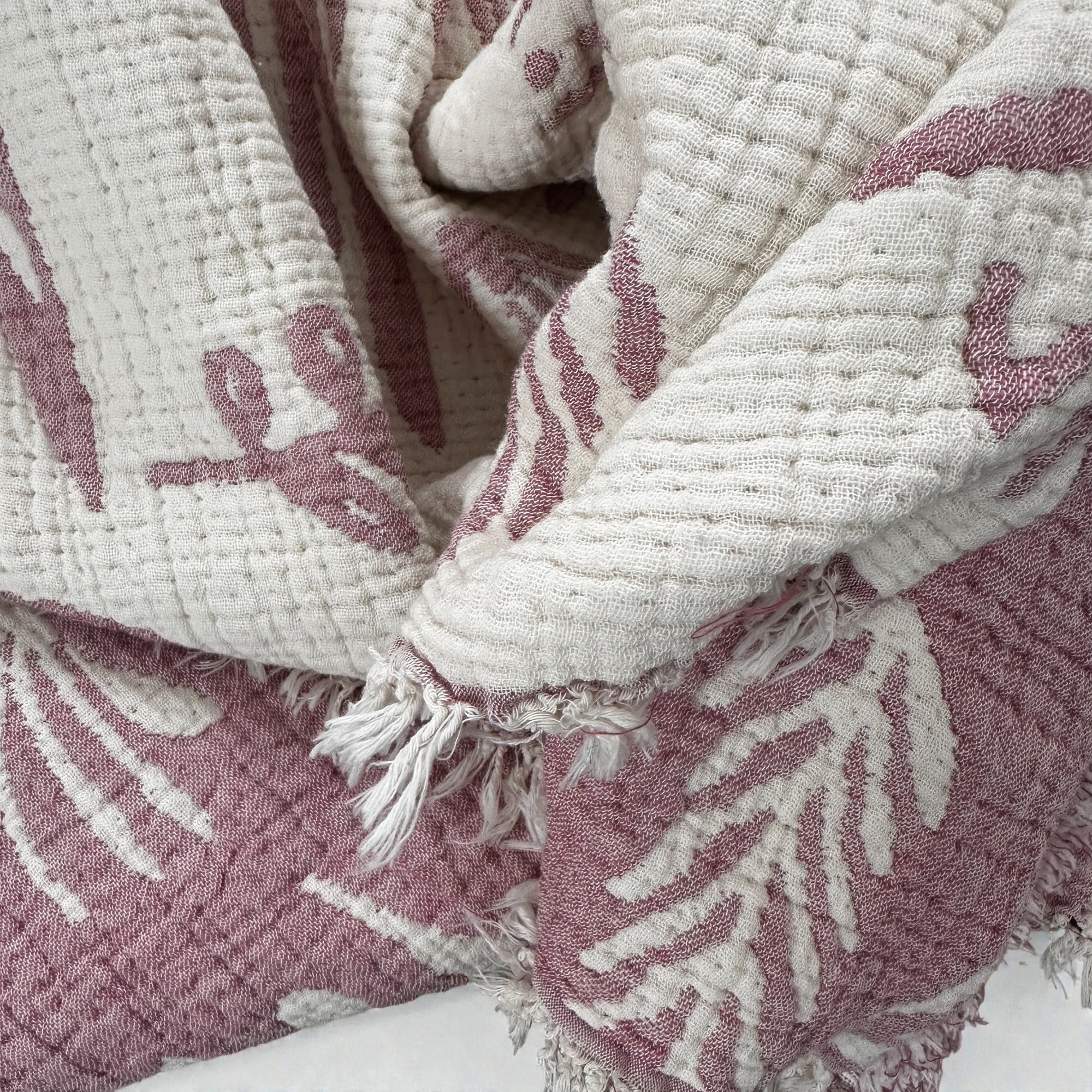Eine Nahaufnahme der LEY! CALMA Musselin Decke in Rosé-Weiß (2,30 x 2,40 m) zeigt ihre abstrakten Muster, das weiche Boho-Design, die ausgefransten Kanten und die luftige, atmungsaktive Qualität.
