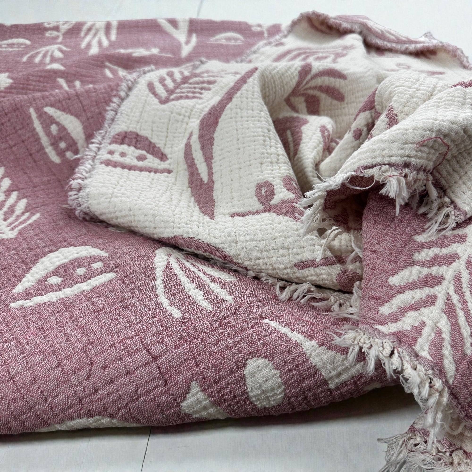 Die CALMA Musselin Decke von LEY! in Rosé-Weiß (2,30 x 2,40 m) besteht aus einem weichen, atmungsaktiven Stoff mit Fransenkanten und einem rosa-weißen, botanischen Boho-Design, das locker gefaltet auf einer hellen Fläche liegt.