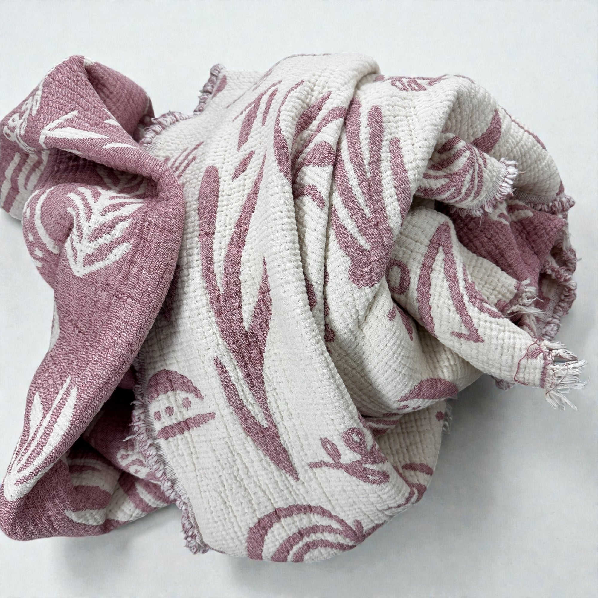 Die CALMA Musselin Decke von LEY! in Rosé-Weiß (2,30 x 2,40 m) zeichnet sich durch eine weiche Textur mit abstrakten botanischen und regenbogenfarbenen Boho-Motiven aus, die auf einem hellen Hintergrund zerknittert dargestellt sind.
