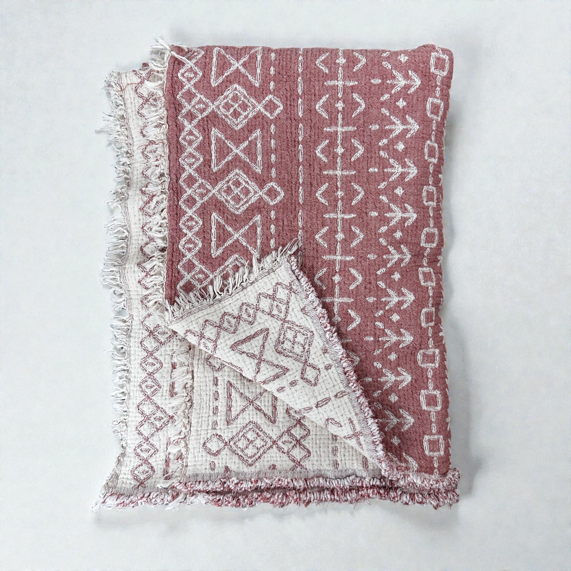 Die CALMA Musselin Decke von LEY! in Rotbraun-Weiß (1,60 x 2,30 m) besticht durch geometrische Muster und Fransenkanten. Gefaltet auf hellem Grund präsentiert, werden das Boho-Design und das atmungsaktive Material durch die sichtbare Rückseite hervorgehoben.