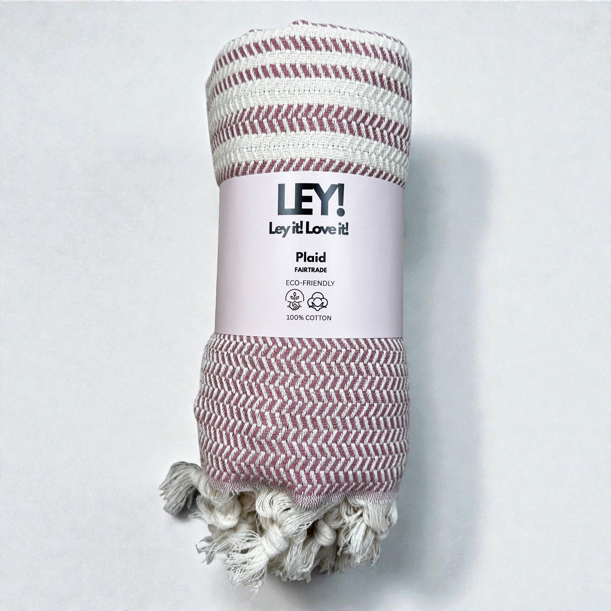 Ein gerolltes, umweltfreundliches LEY! Plaid - Zickzack - Aubergine aus 100% Baumwolle mit rosa-weißem Zickzack-Muster und Fransen an den Enden - ideal als Tagesdecke oder Picknickdecke - eingewickelt in ein Etikett mit der Marke LEY! und Produktinformation.