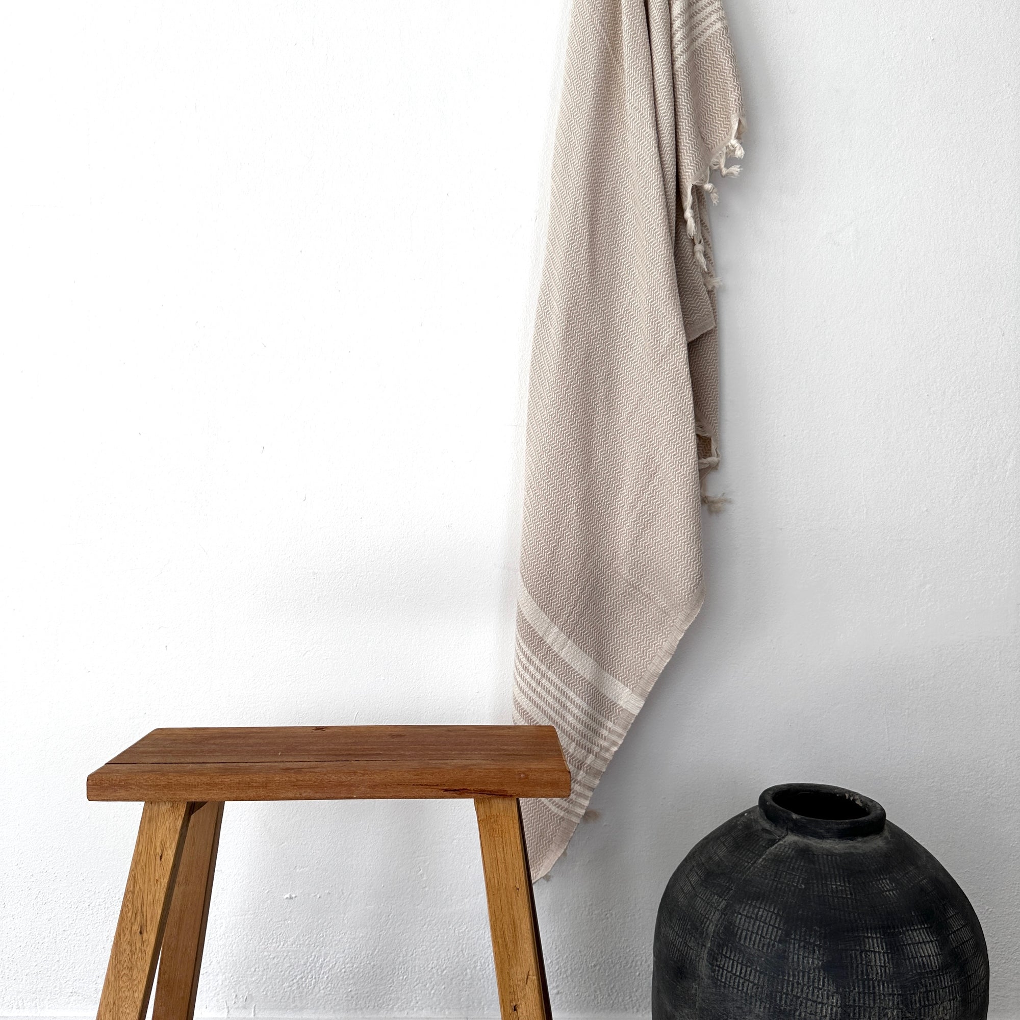 Ein Holzhocker steht neben einer dunklen, runden Keramikvase auf dem Boden, darüber ist das LEY! Plaid - Zickzack - Beige von LEY! an einer weißen Wand darüber drapiert. Das Arrangement schafft einen minimalistischen, aufgeräumten Look.