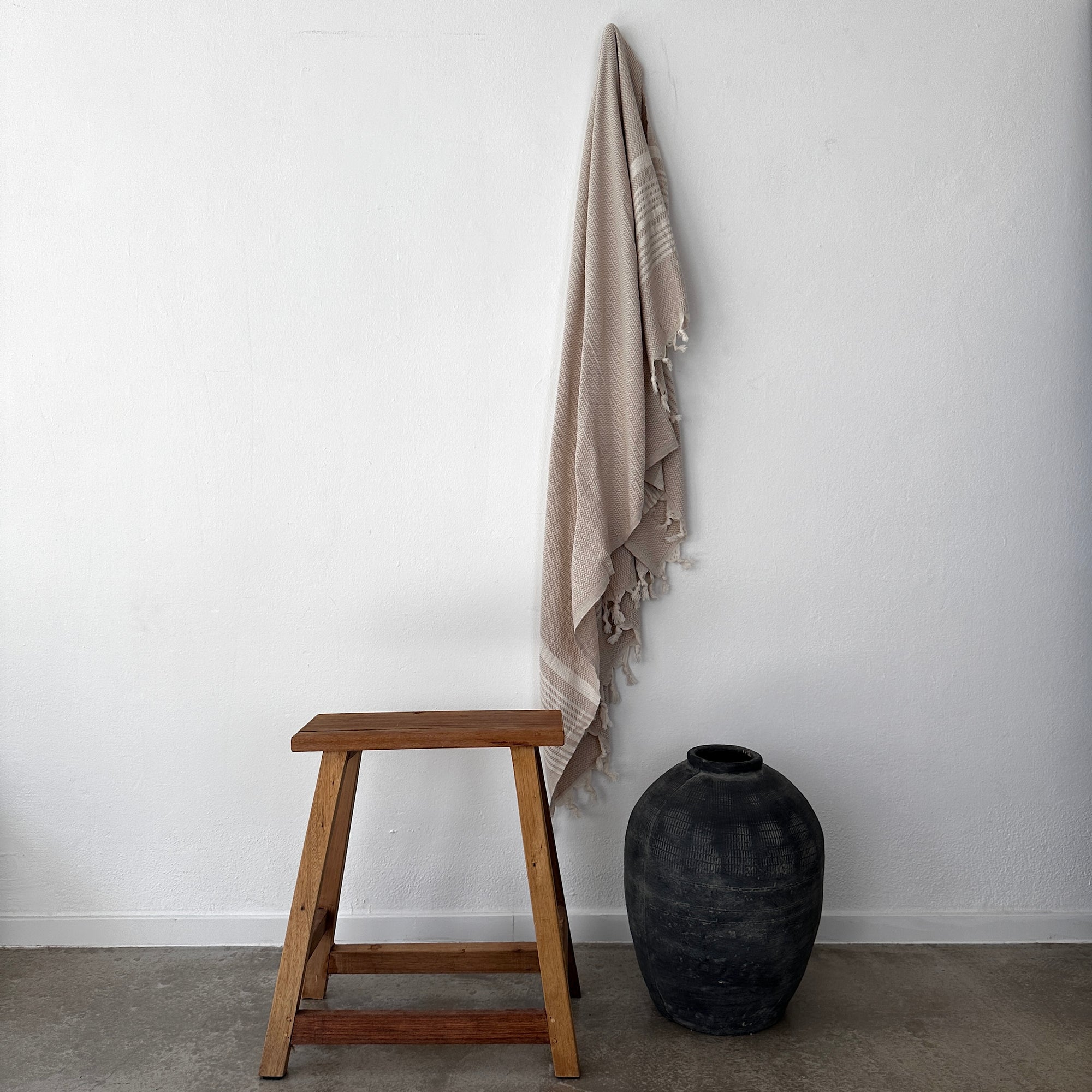 Eine minimalistische Szene mit einem Holzhocker, einer schwarzen Keramikvase auf dem Boden und dem LEY! Plaid - Zickzack - Beige von LEY! an einer weißen Wand hängt.