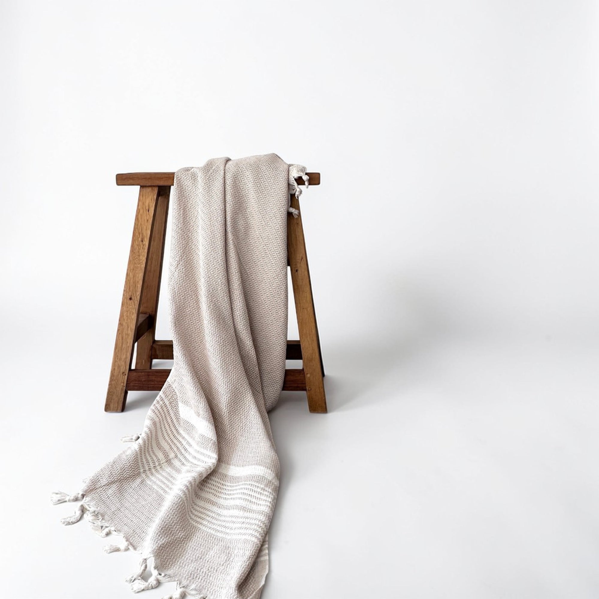 Die LEY! Plaid - Zickzack - Beige gewebte Decke von LEY! mit Quasten ist über einen schlichten Holzhocker drapiert und steht vor einem schlichten weißen Hintergrund.