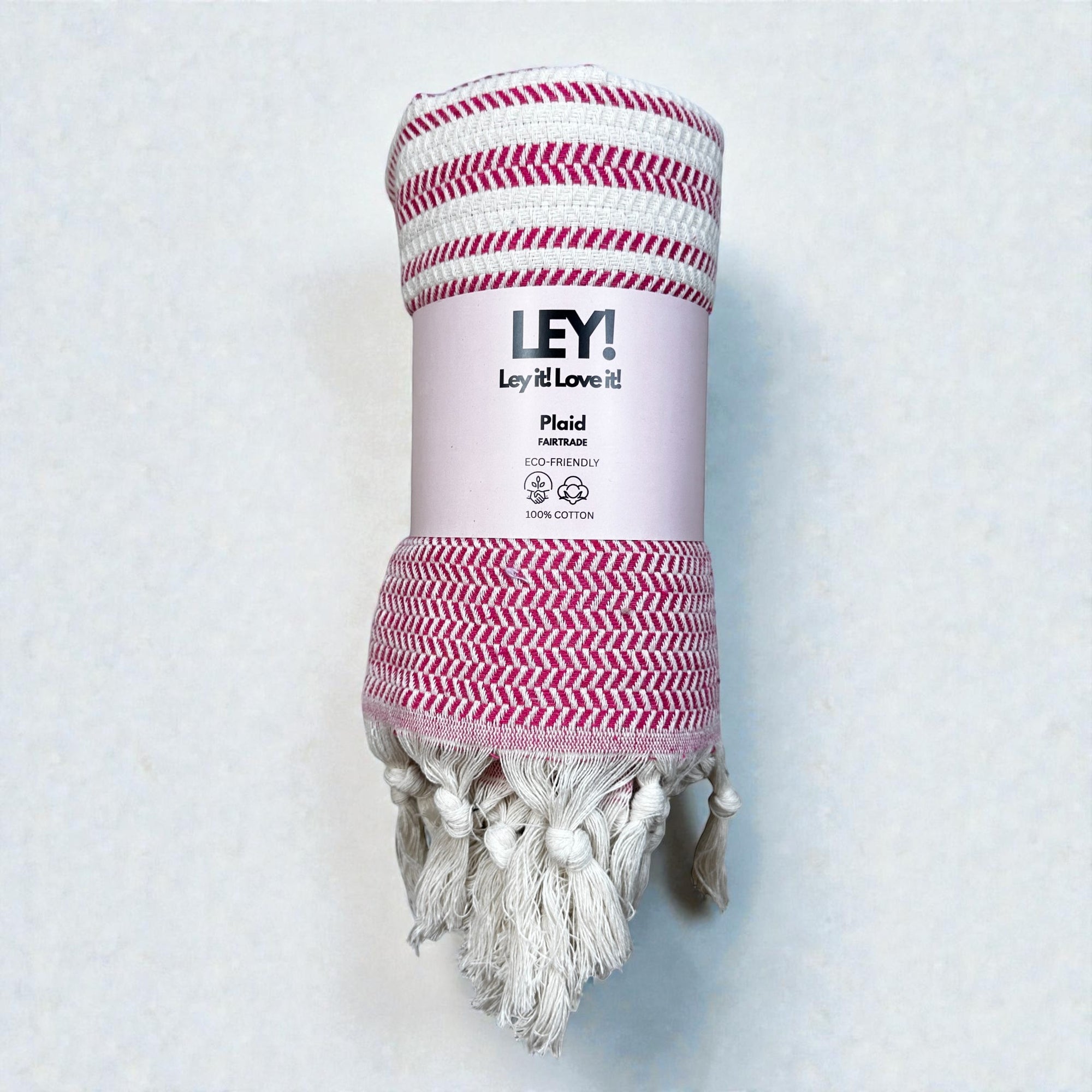 Eine aufgerollte rosa-weiße, zickzackgemusterte LEY! Plaid - Zickzack - Rosa Baumwolldecke mit Quasten, beschriftet mit "LEY! Plaid, Eco-Friendly, 100% Baumwolle", ist auf einem hellen Hintergrund abgebildet.